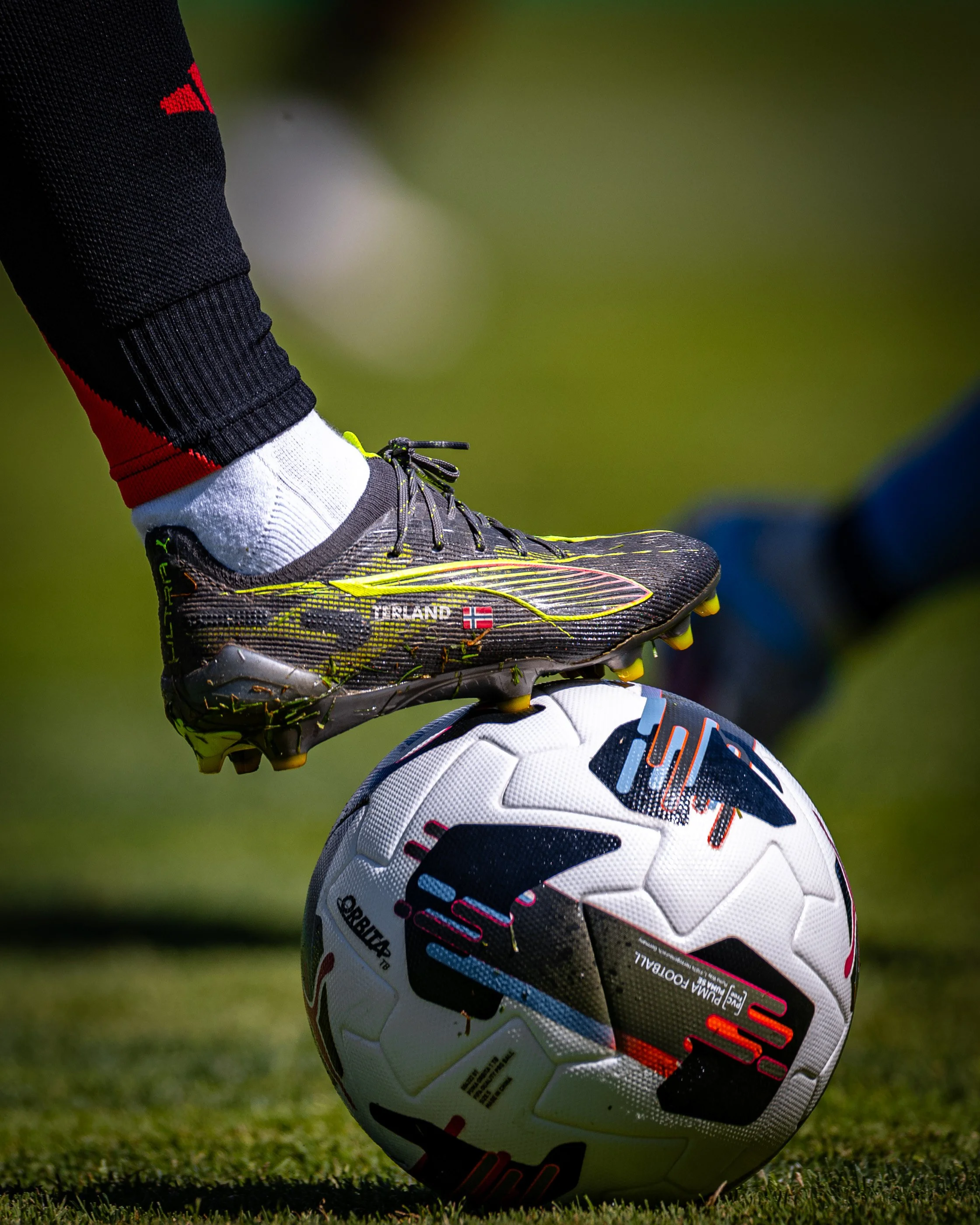 Close-up of a black, white, and colored soccer ball on grass with a partially visible athlete's foot in a gray and black soccer cleat, wearing a black sock with red accents.