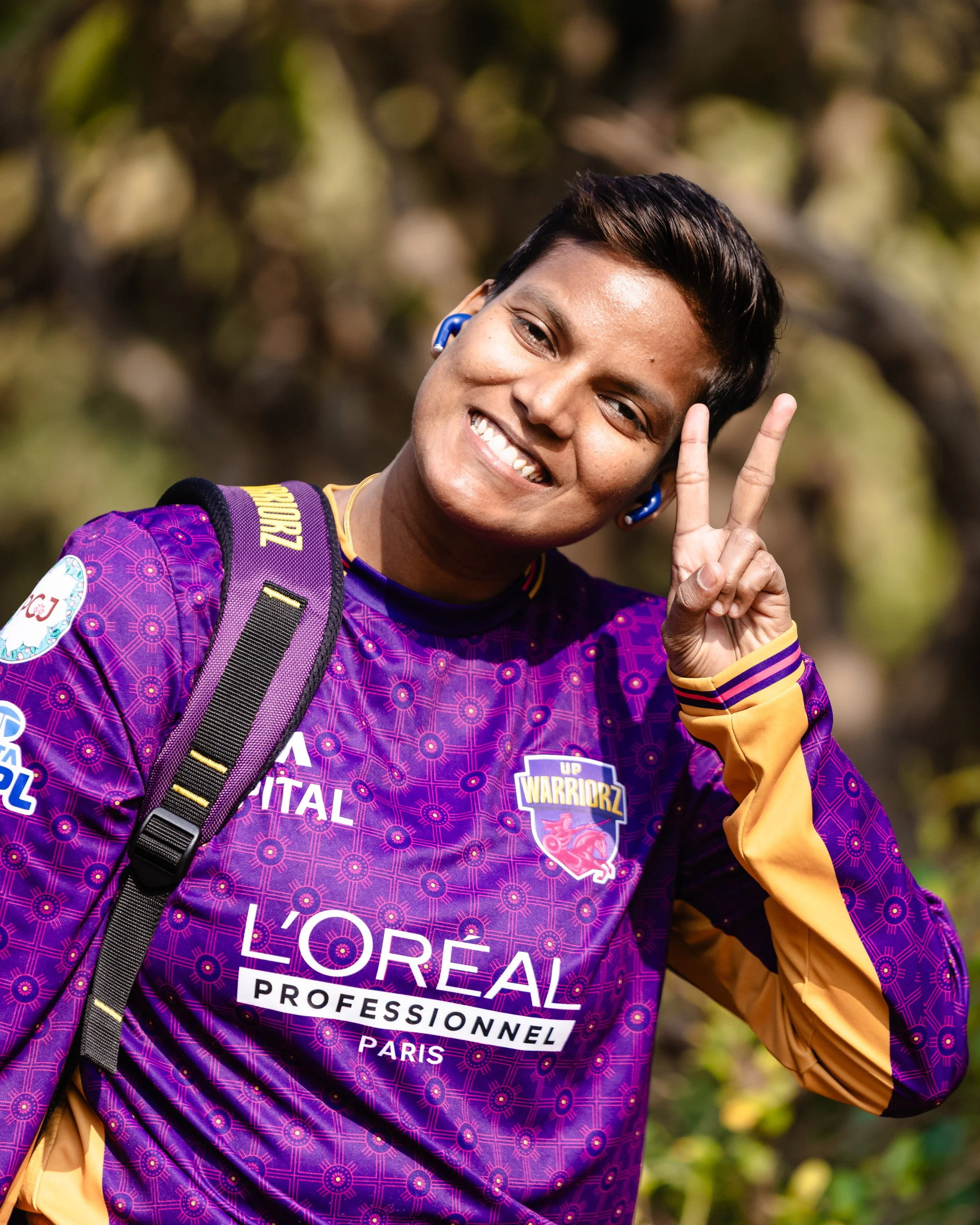 A young woman outdoors, smiling and making a peace sign with her right hand. She is wearing a purple sports jersey with yellow accents, a backpack, and blue earbuds. The background is blurred with trees and sunlight.