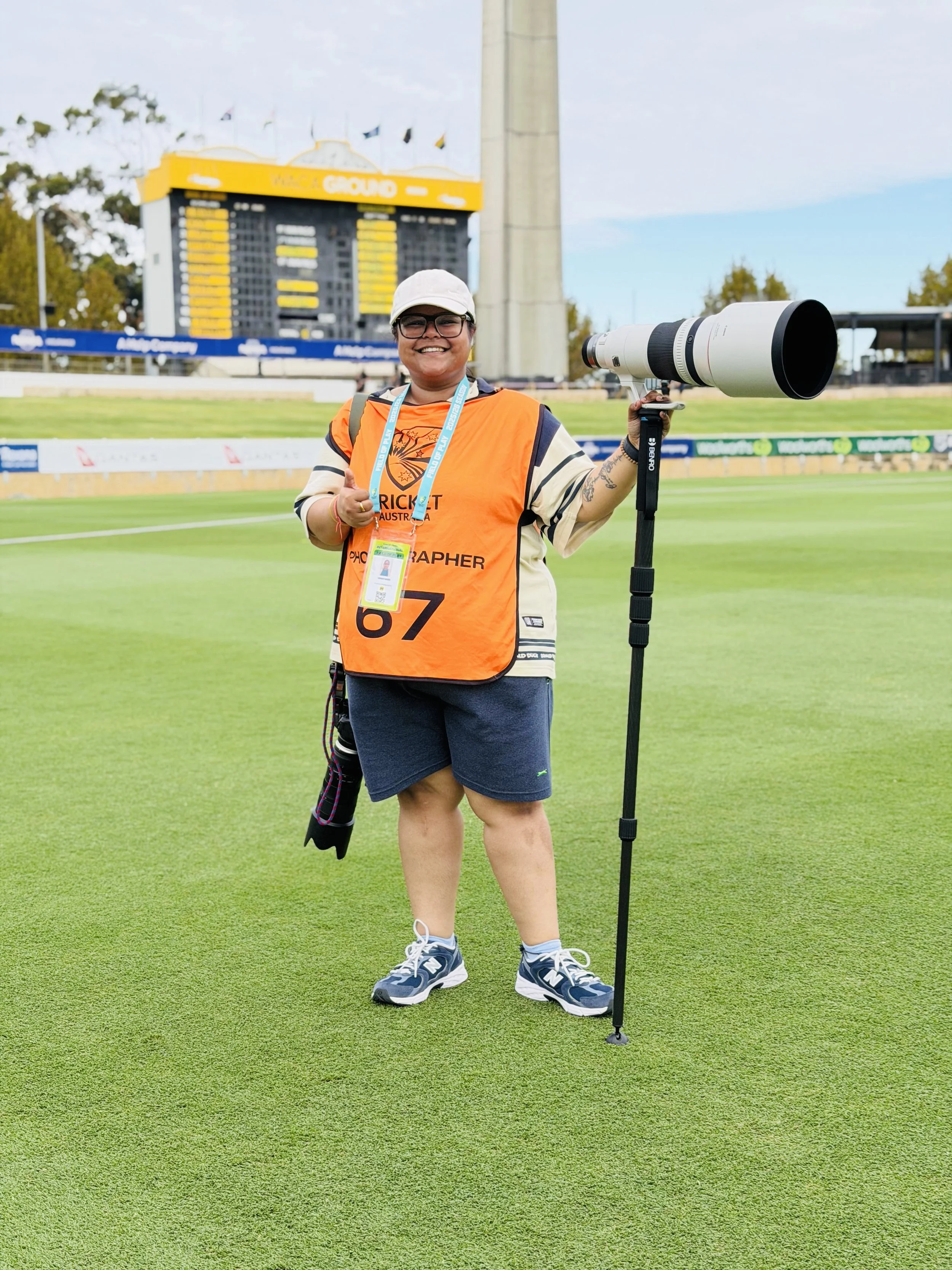 A person smiling and giving a thumbs up on a cricket field, holding a large camera with a telephoto lens, wearing a white cap, glasses, an orange vest, shorts, and sneakers, with a scoreboard and scoreboard tower in the background.