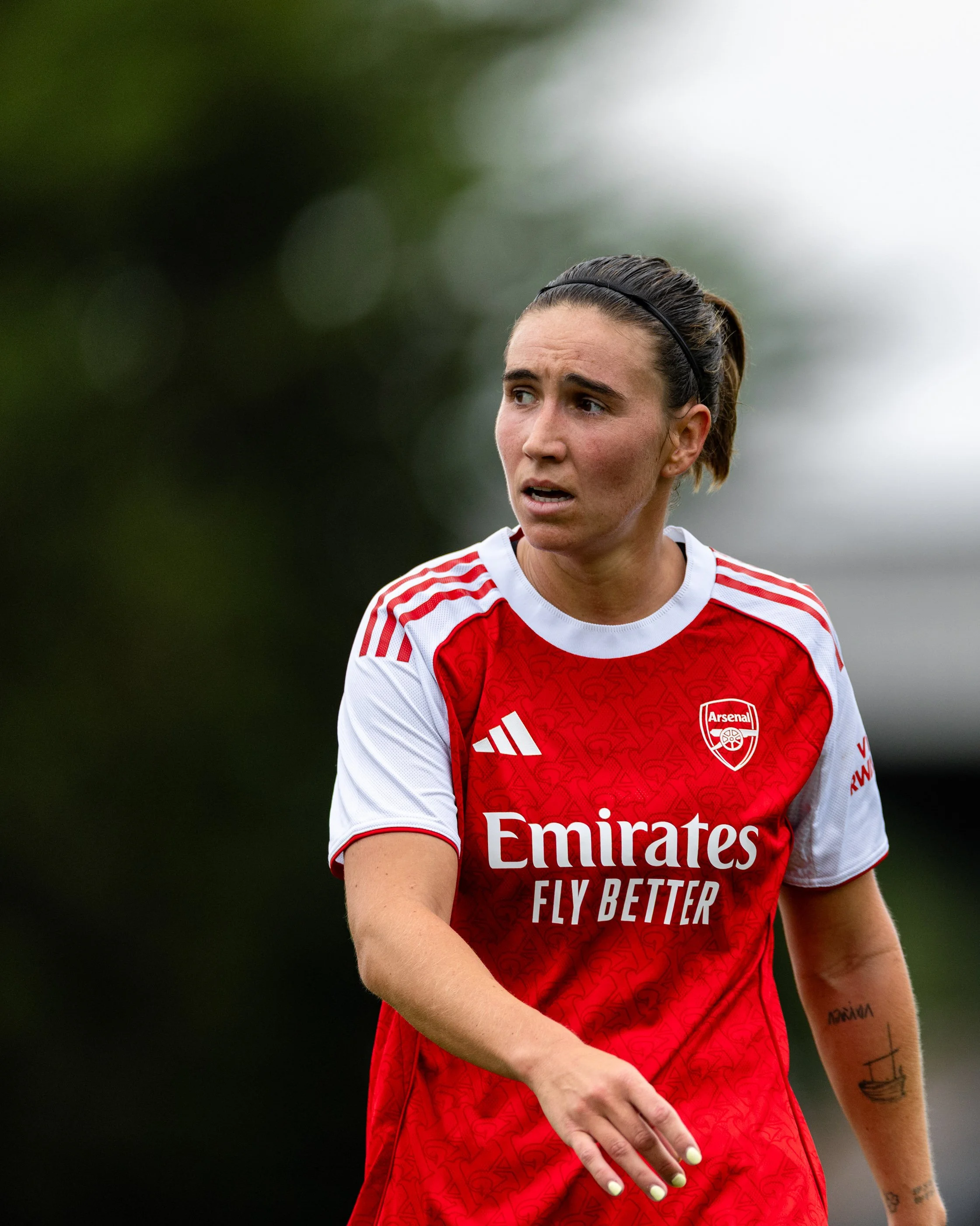 A female soccer player wearing an Arsenal football club jersey with a red and white design, standing outdoors on a cloudy day, with blurred greenery in the background.
