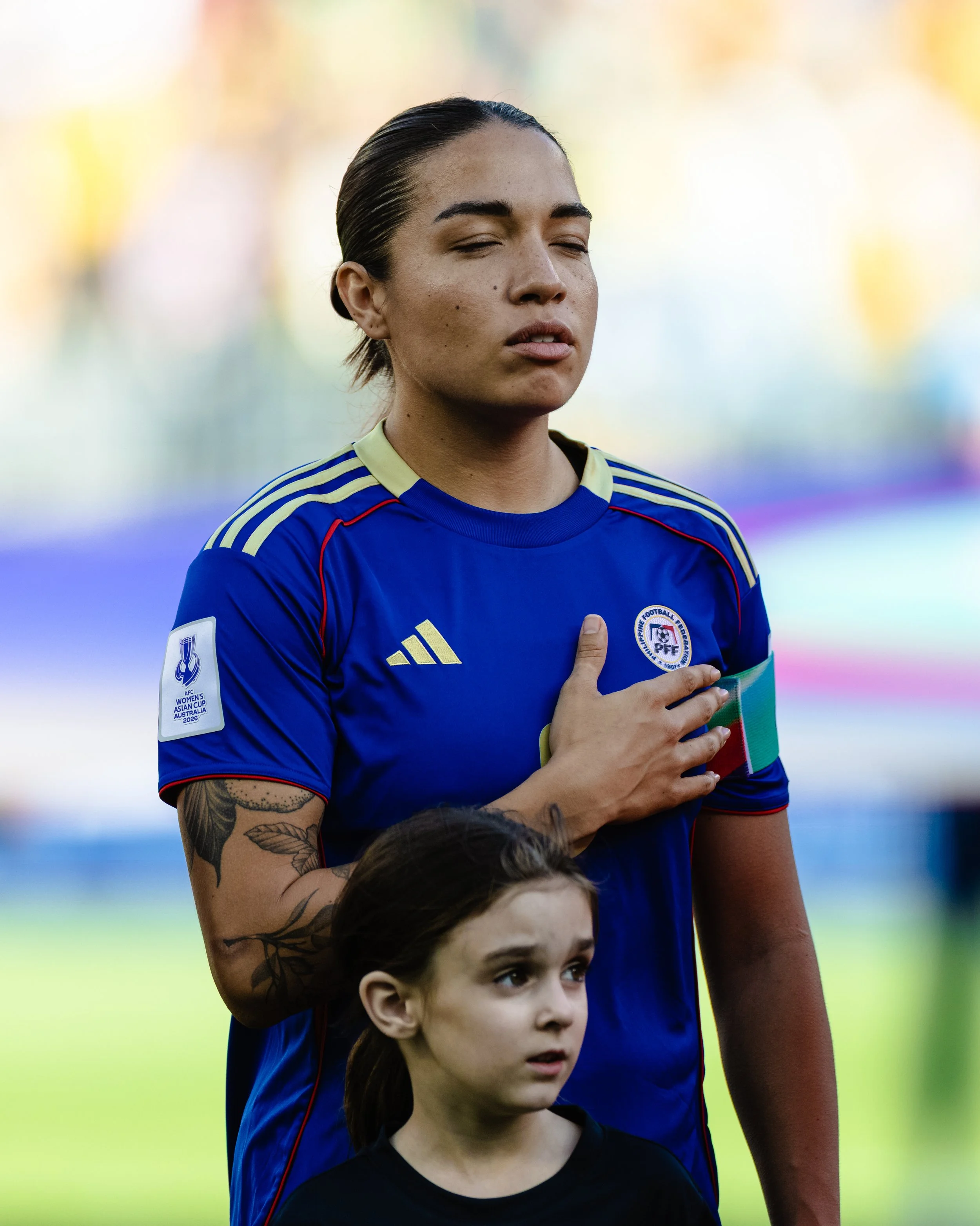A female soccer player standing with a young girl during the national anthem, with her hand over her heart, wearing a blue jersey with Philippines and AFC Women's Asian Cup logos.