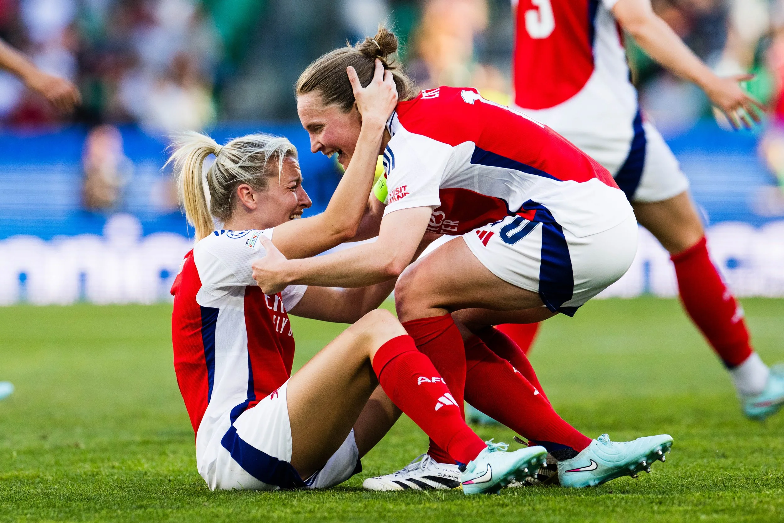 Two female soccer players in red and white uniforms celebrate on the field, with one sitting on the ground and the other crouching and holding her head, both smiling joyfully.