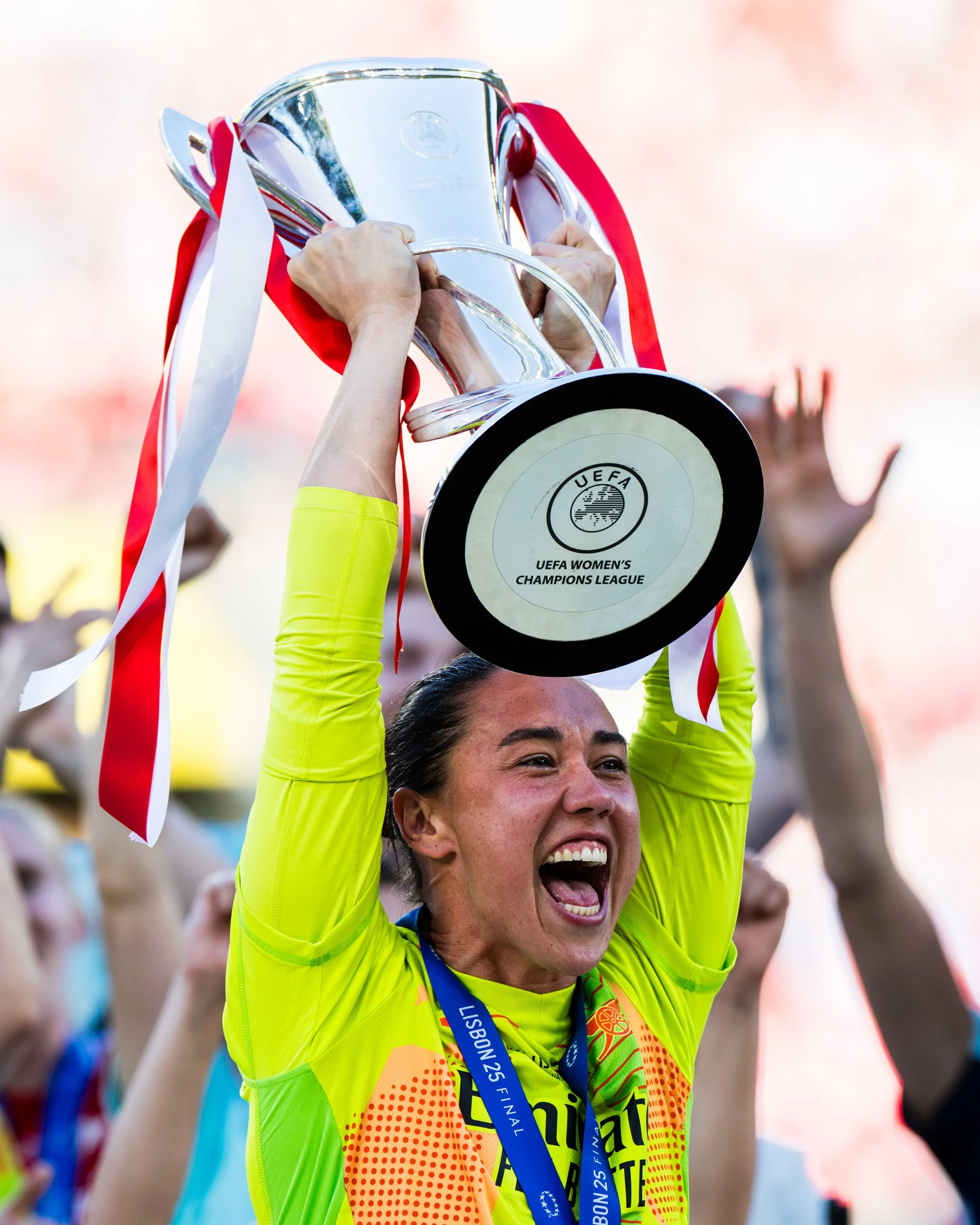 A woman holding a shiny trophy above her head in celebration, wearing a bright yellow and orange sports jersey with a medal around her neck, at a sports event with other people in the background.