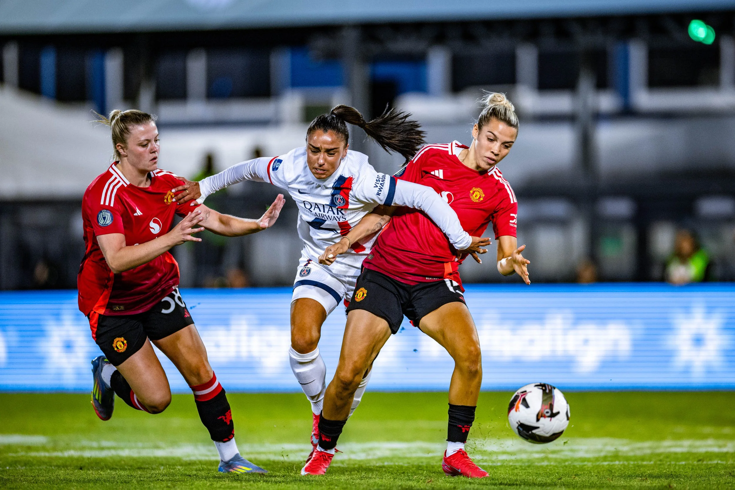 Three women playing soccer on a field during a match, two of them wearing Manchester United jerseys and the third in a Paris Saint-Germain jersey, all vying for the ball.