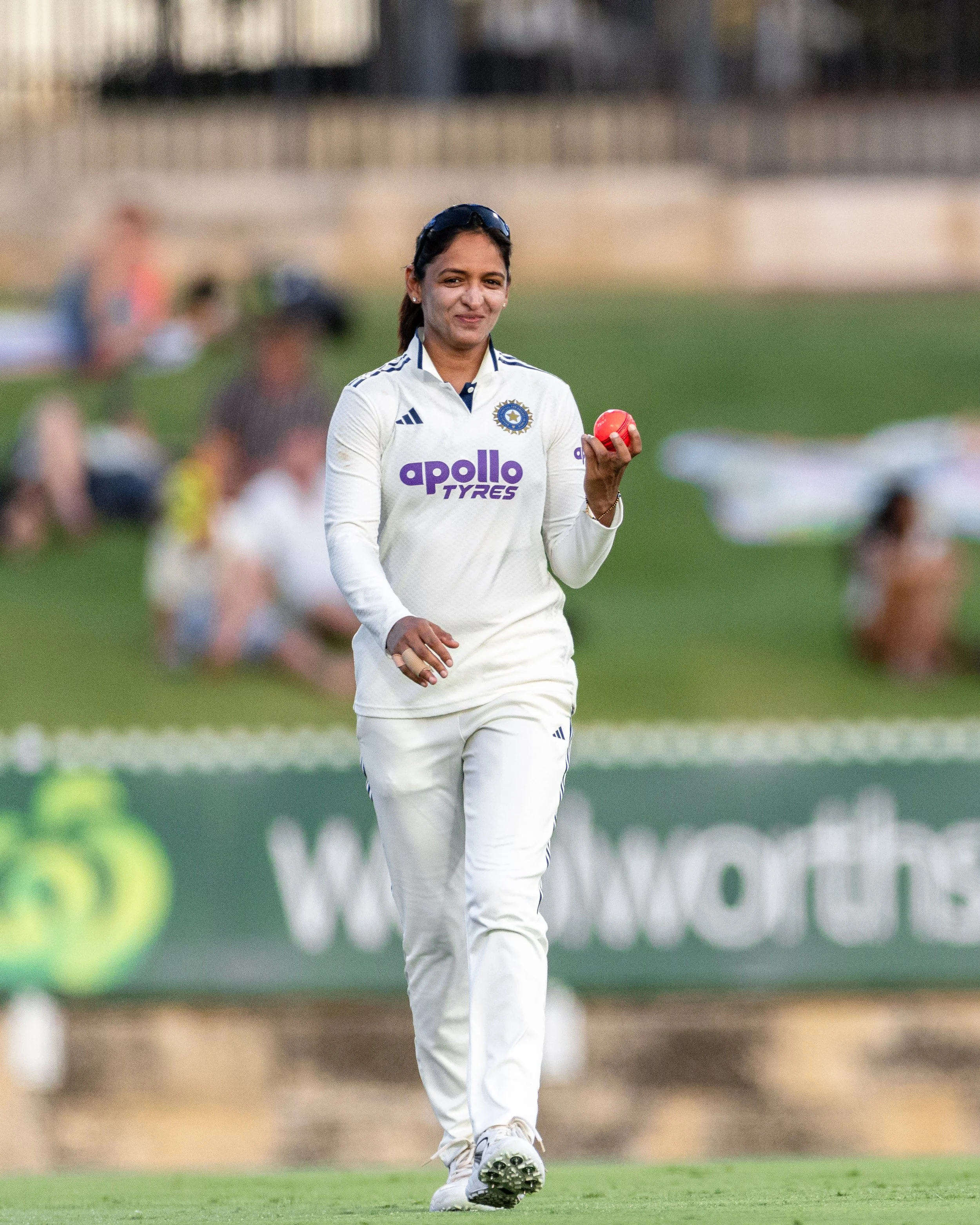 A female cricket player in white uniform holding a red cricket ball on a cricket field.
