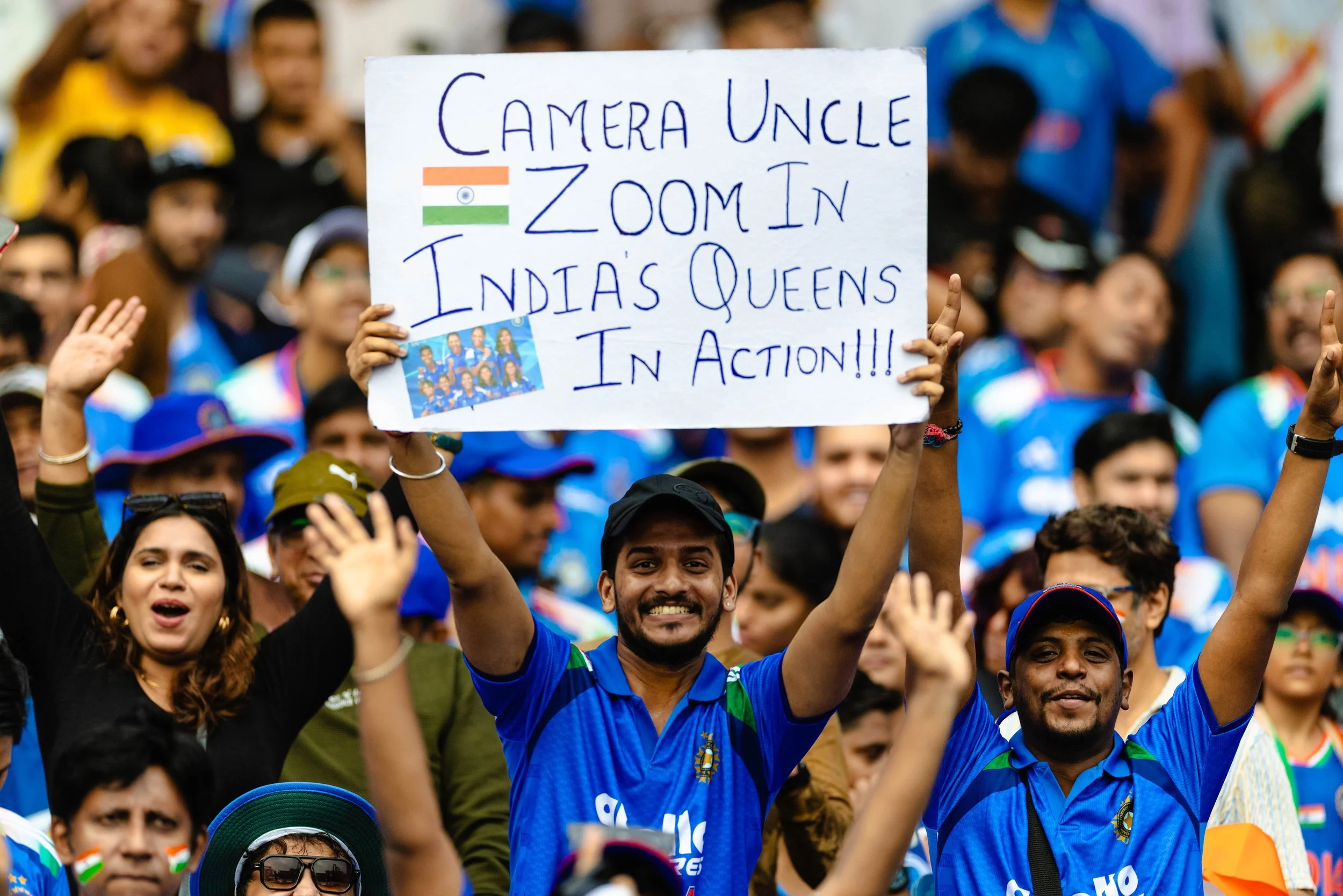 Crowd of people at a sporting event wearing blue India jerseys, with a man holding a sign that reads, "Camera Uncle zoom in India’s Queens in action!!!"