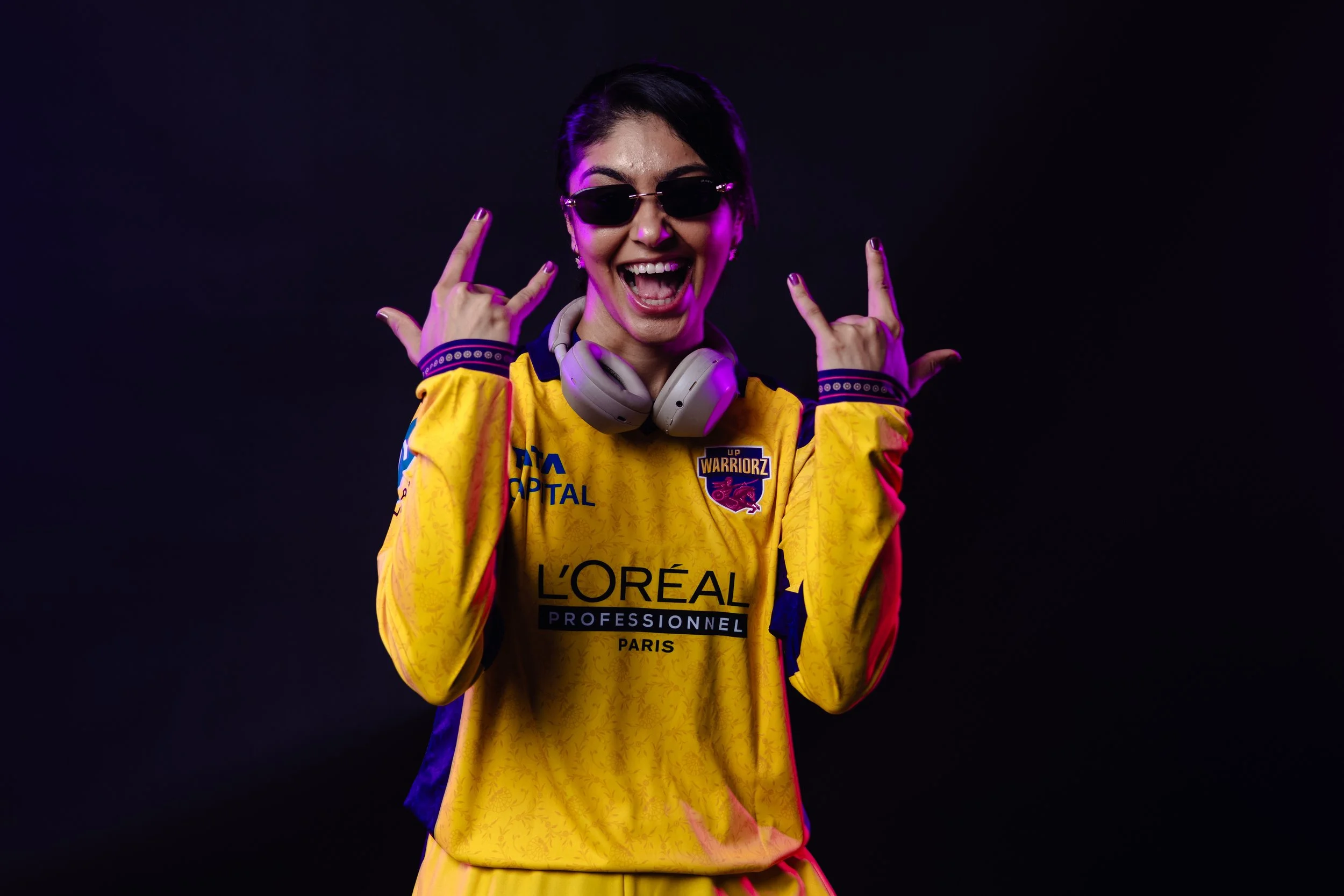 A woman in a yellow sports jersey with sunglasses and headphones making rock hand gestures and smiling against a dark background.