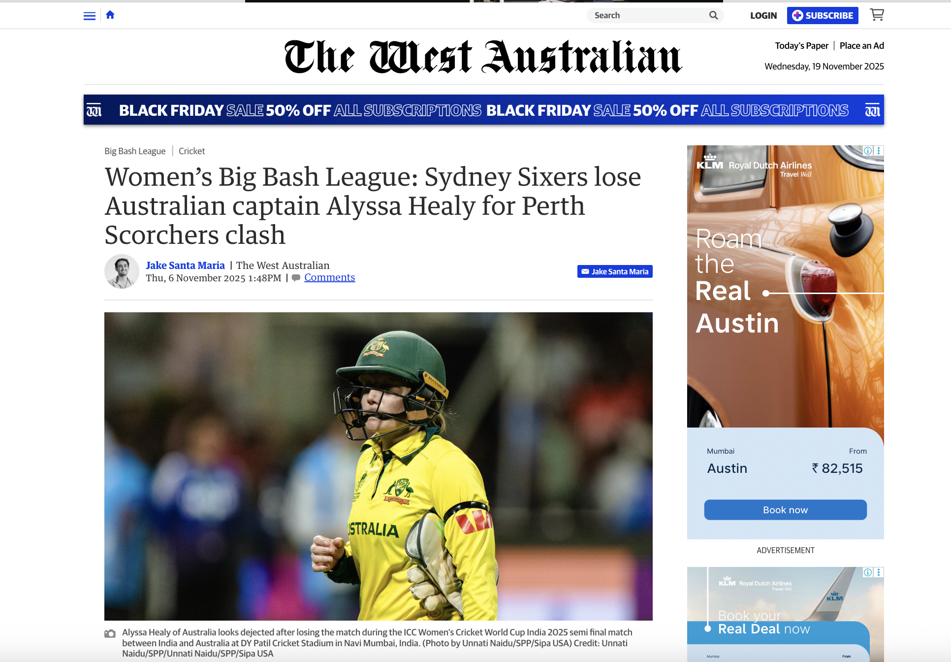 A female Australian cricket player in yellow team uniform and helmet on a cricket field. She is holding cricket pads and a glove, with other players blurred in the background.