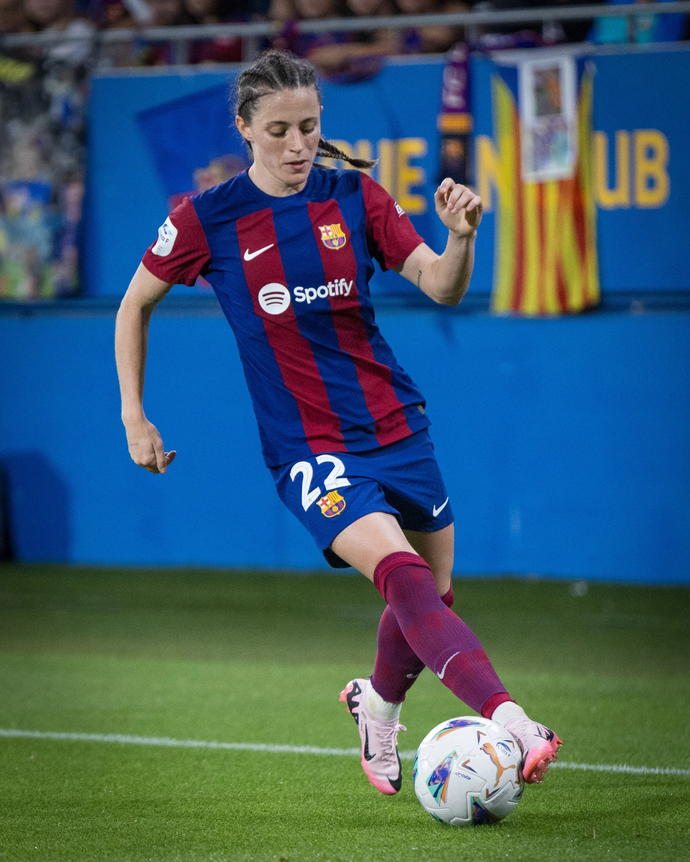 A female soccer player wearing a FC Barcelona uniform is about to kick a soccer ball on the field. She has cornrow braids and is wearing pink and white cleats, with a background of a blue wall and a Catalonia flag.