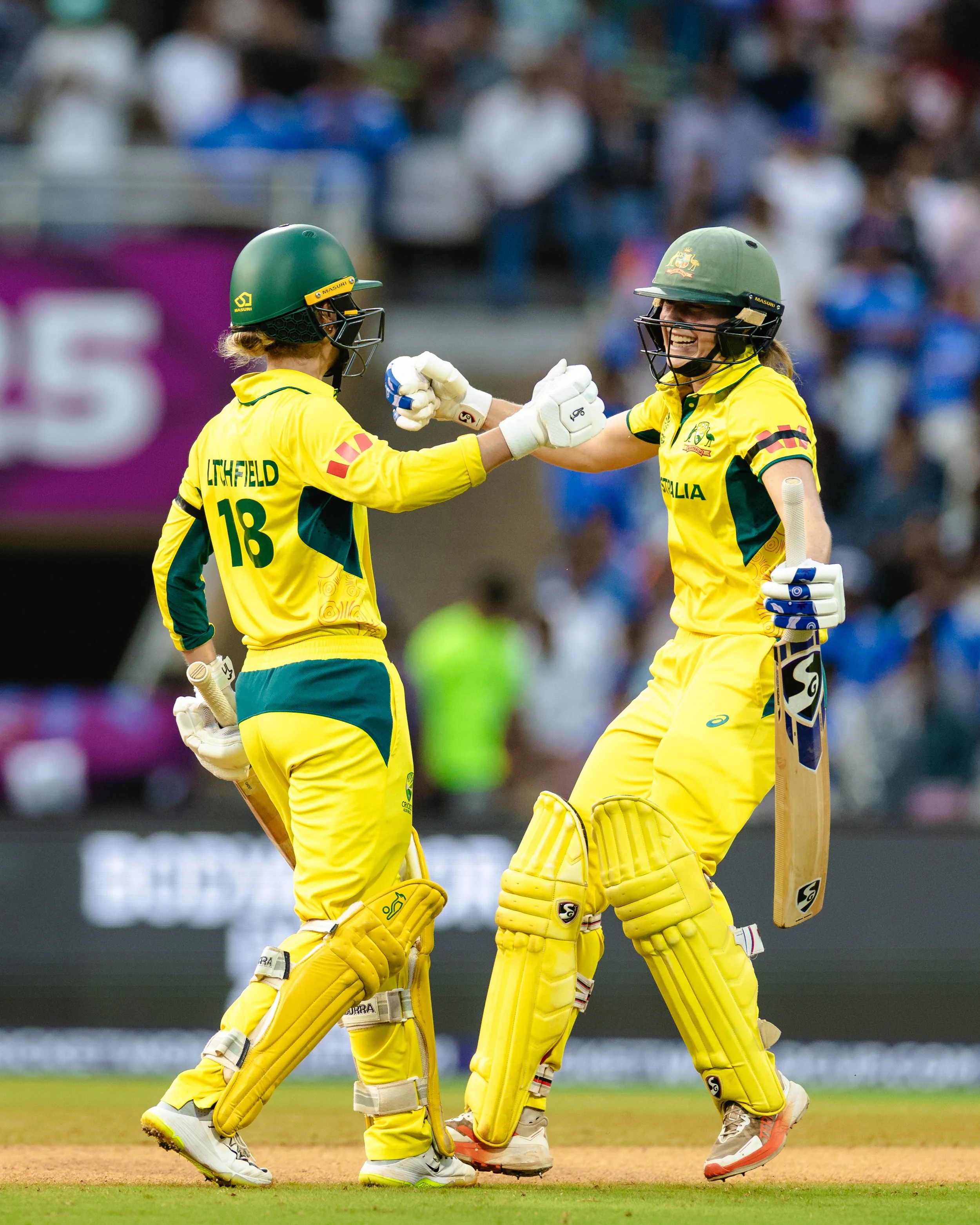 Two female cricketers in yellow uniforms celebrating on the cricket field, one with a helmet with Australia emblem, and the other with a green helmet, both with batting gloves and pads. They are fist bumping.