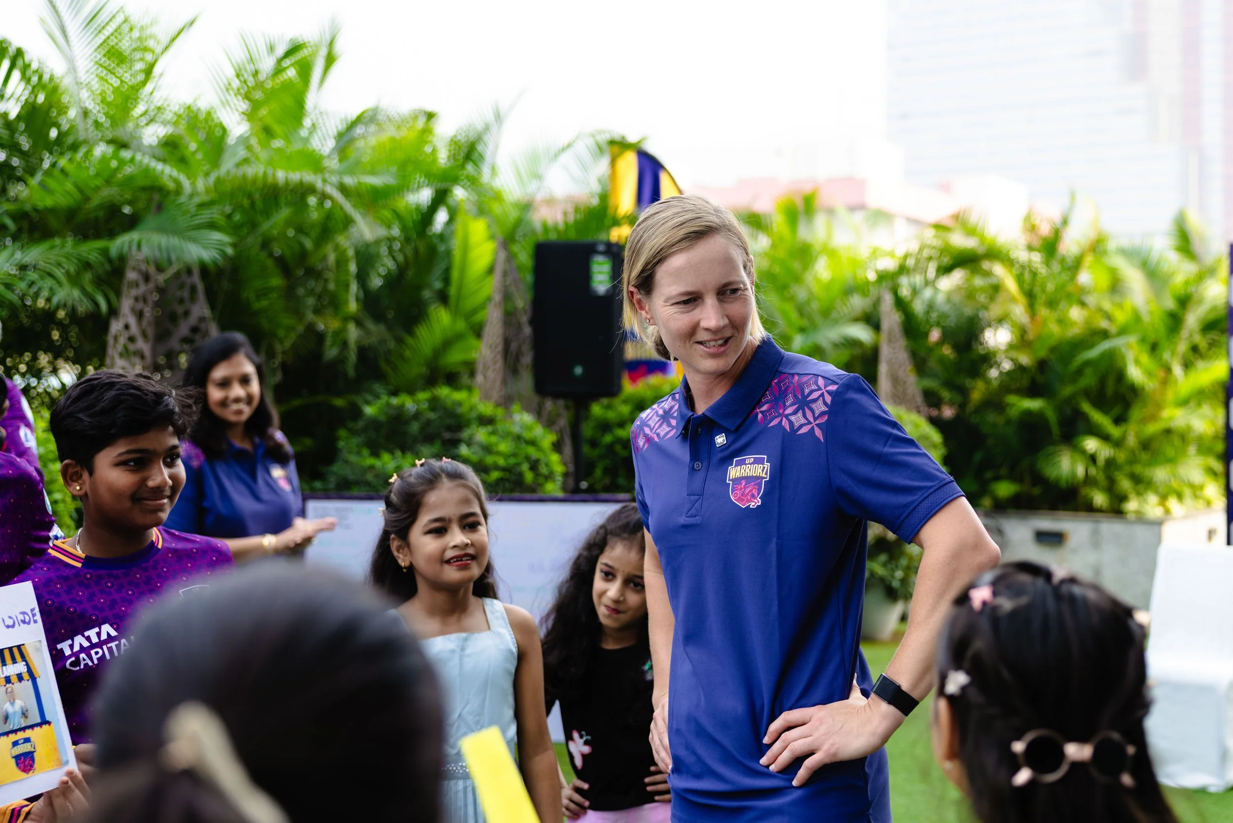 A woman with short blonde hair wearing a blue sports shirt is surrounded by children of diverse ethnicities, outdoors with lush green plants and buildings in the background.