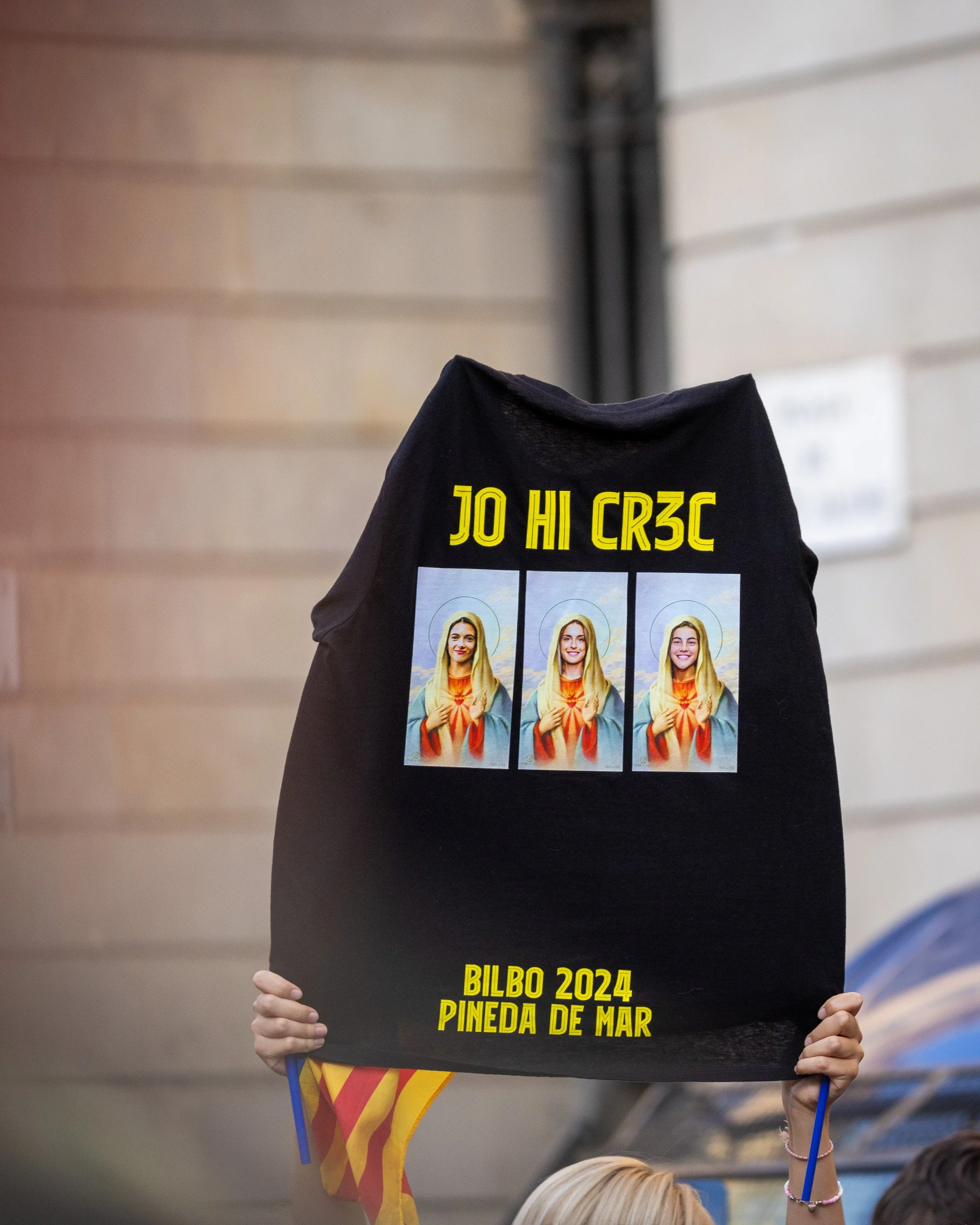 A person holds a sign with images of a woman dressed as Jesus, along with the text 'JO HI CRC JO CR3C' and 'BILBO 2024 DINO 2024 PINEDA DE MAR.' Another person holds a Catalan flag in the background.