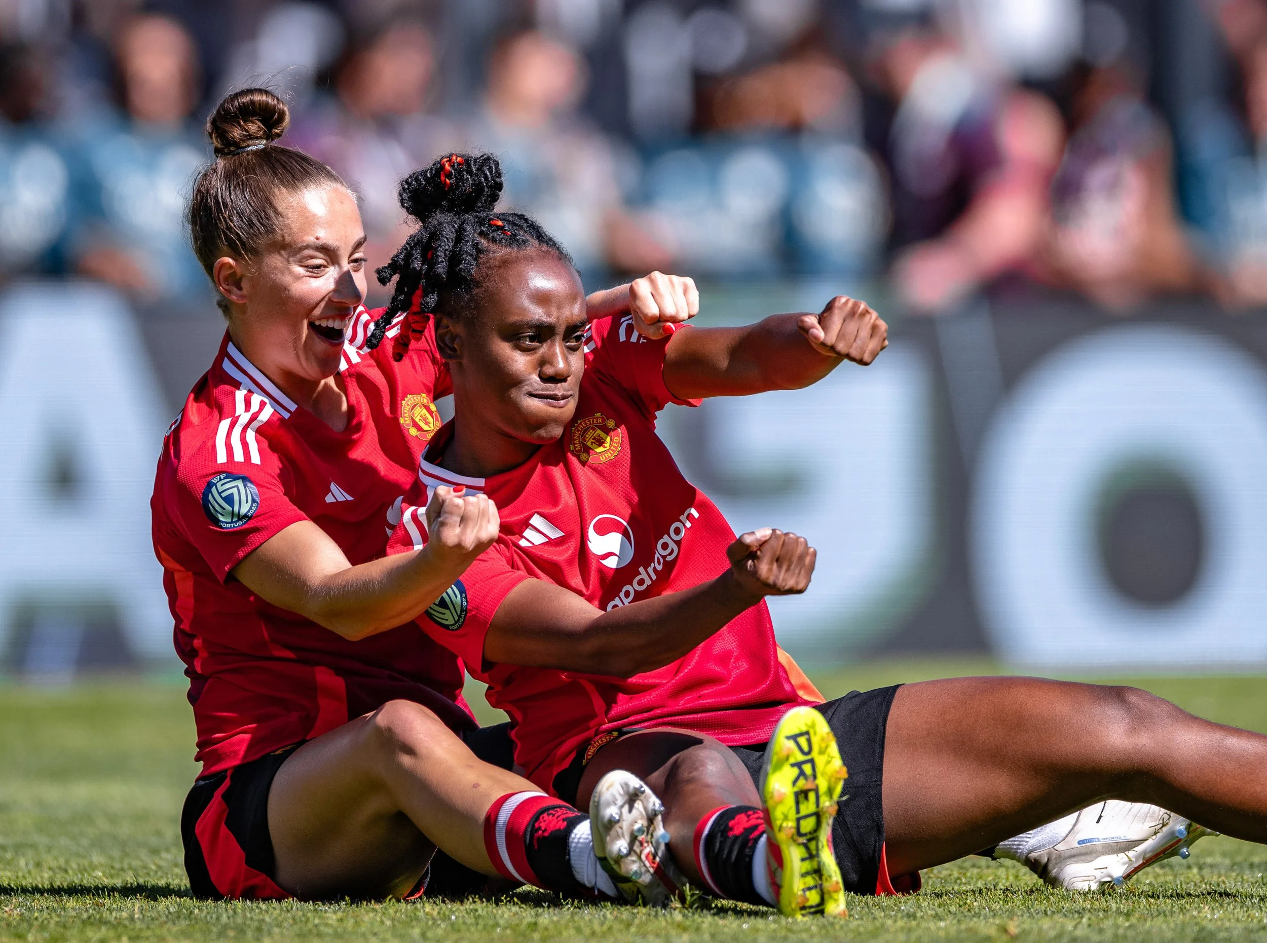 Two female soccer players in red jerseys celebrating on the field, sitting on the grass, with blurred spectators in the background.