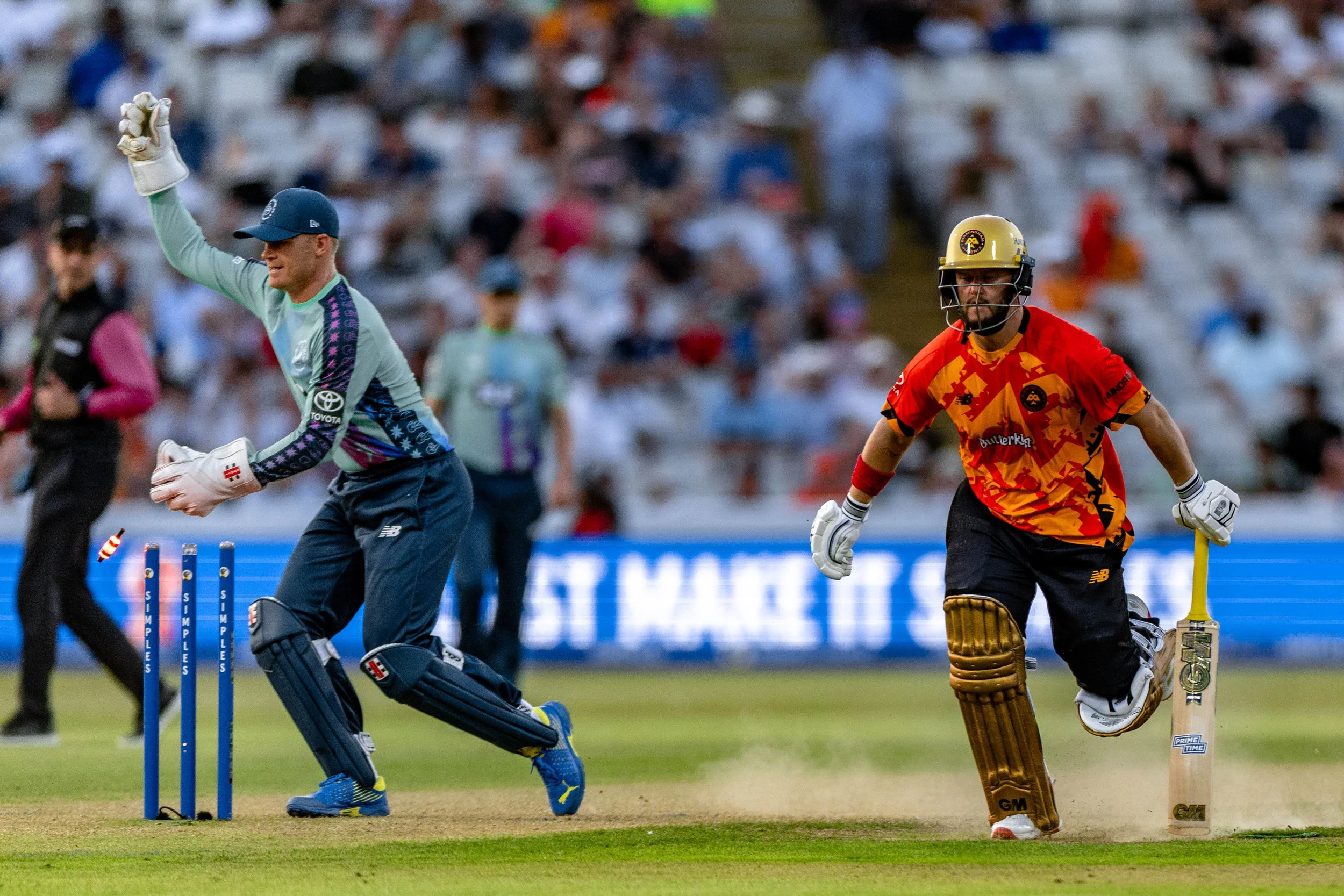 Cricket players on the field during a match, with a crowd in the stands. The player in the foreground is running past the stumps while another player is kneeling behind him.
