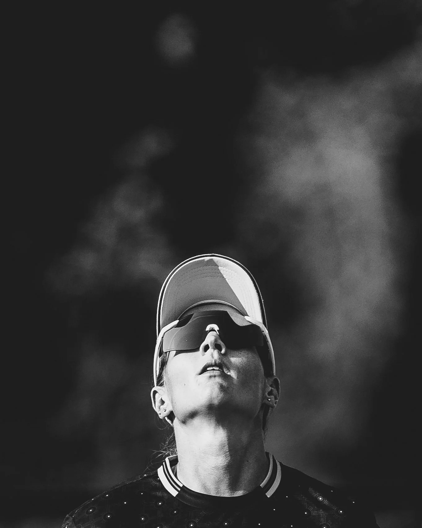 A woman with sunglasses and a baseball cap looking upward, with a cloudy sky in the background.