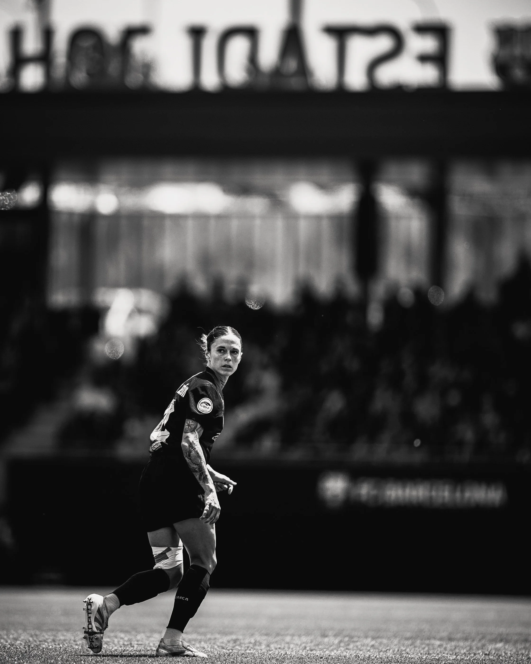 Black and white photo of a female soccer player in motion on a field, with a blurred stadium background.