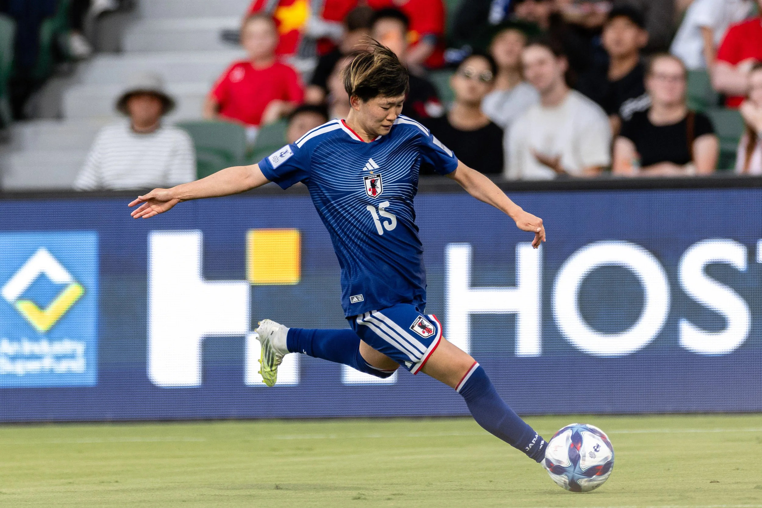 A female soccer player wearing a blue uniform with the number 15 is kicking a black and white soccer ball on the field during a match. Spectators are visible in the background.