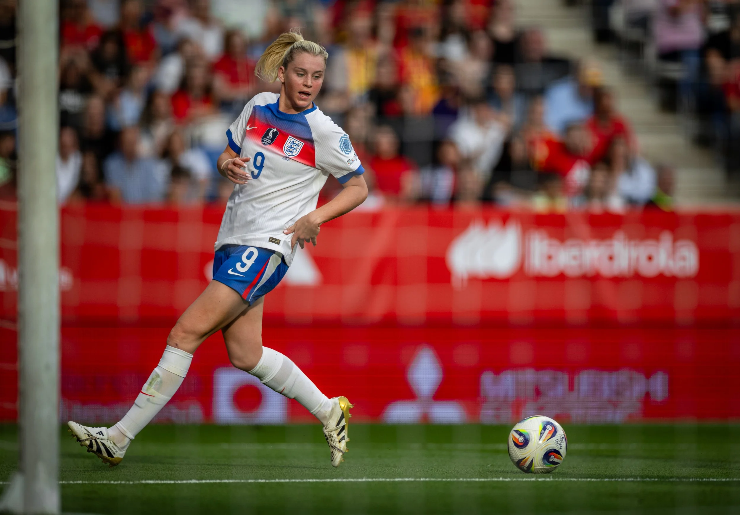 A female soccer player in an England national team jersey runs towards the goal during a match, near the goalpost with a blurred audience in the background.