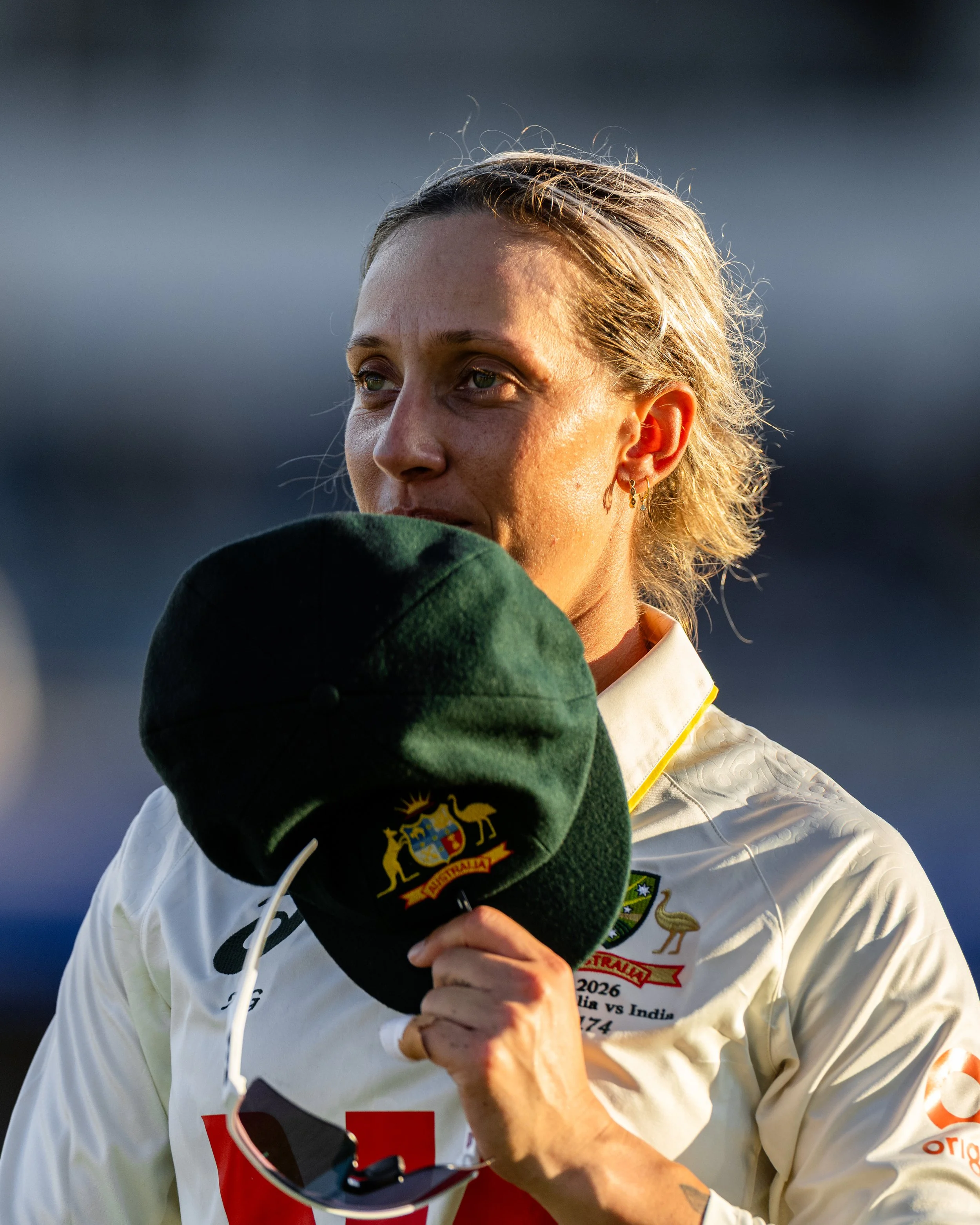 Close-up of a woman with blonde hair, wearing a white sports jacket with Australian flags and text, holding a black hat with an Australian emblem over her face, in a sports context.