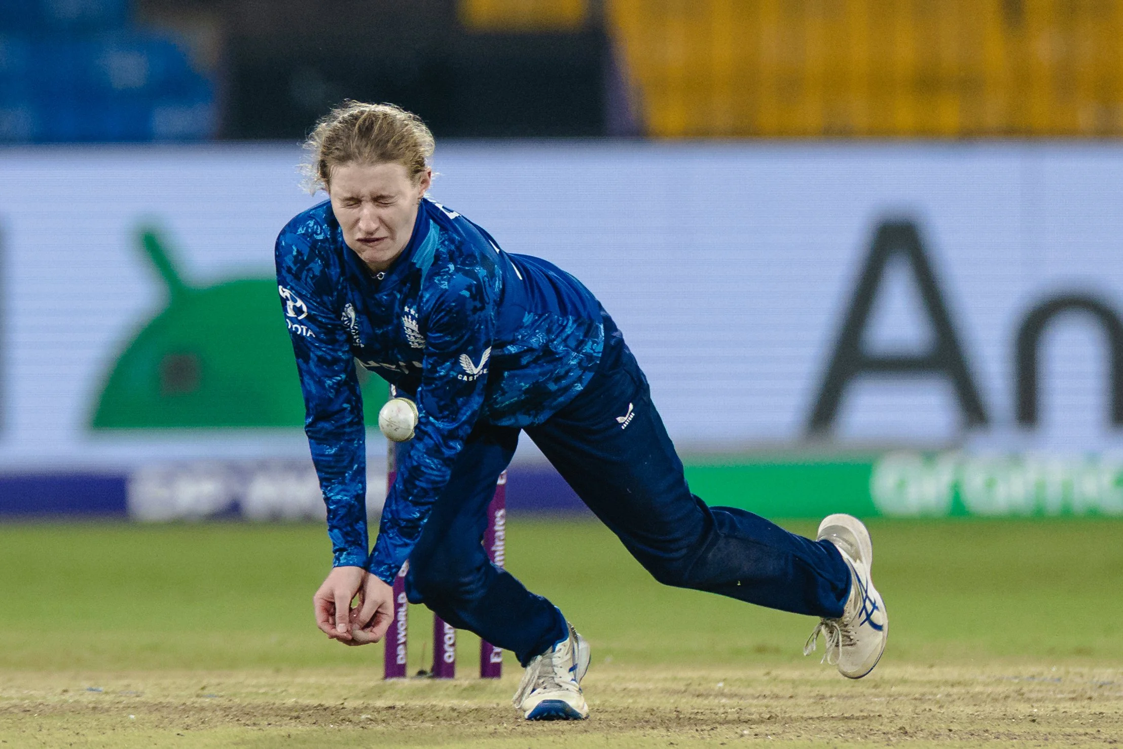 A female athlete in a blue sports uniform is kneeling on a cricket pitch, appearing to be upset or exhausted after a game.