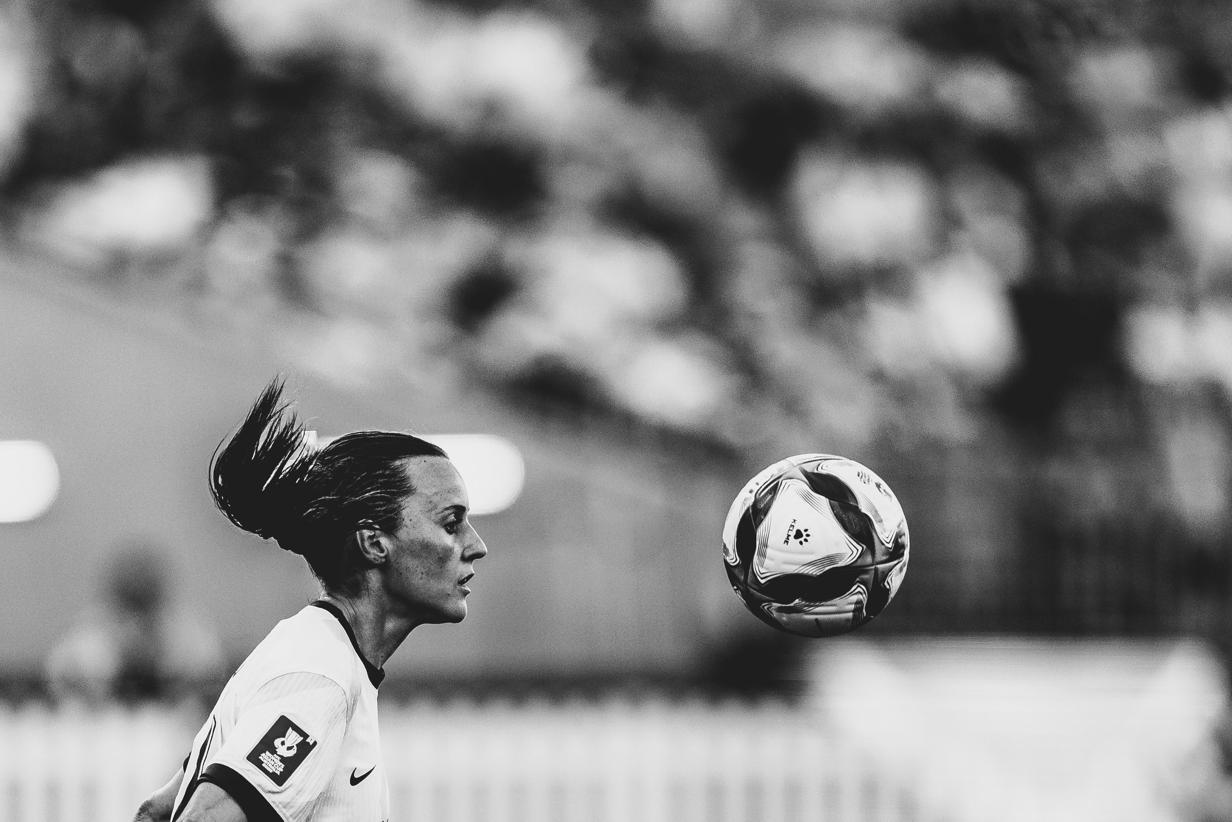 A female soccer player with dark hair tied back, wearing a white uniform, is focusing on a soccer ball in mid-air during a game.