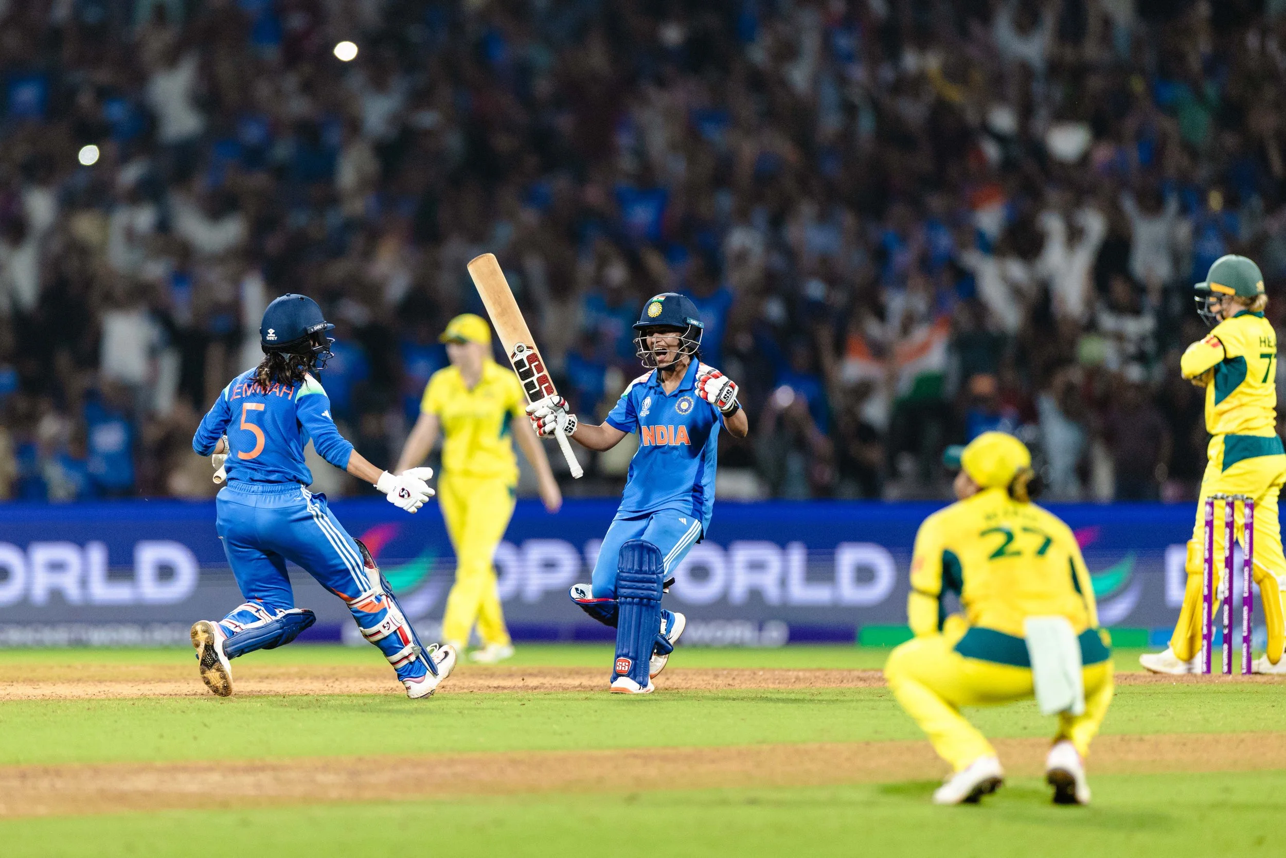 Cricket players celebrating on the field during a match. The Indian player is holding a bat and smiling, while other players and the crowd are visible in the background.