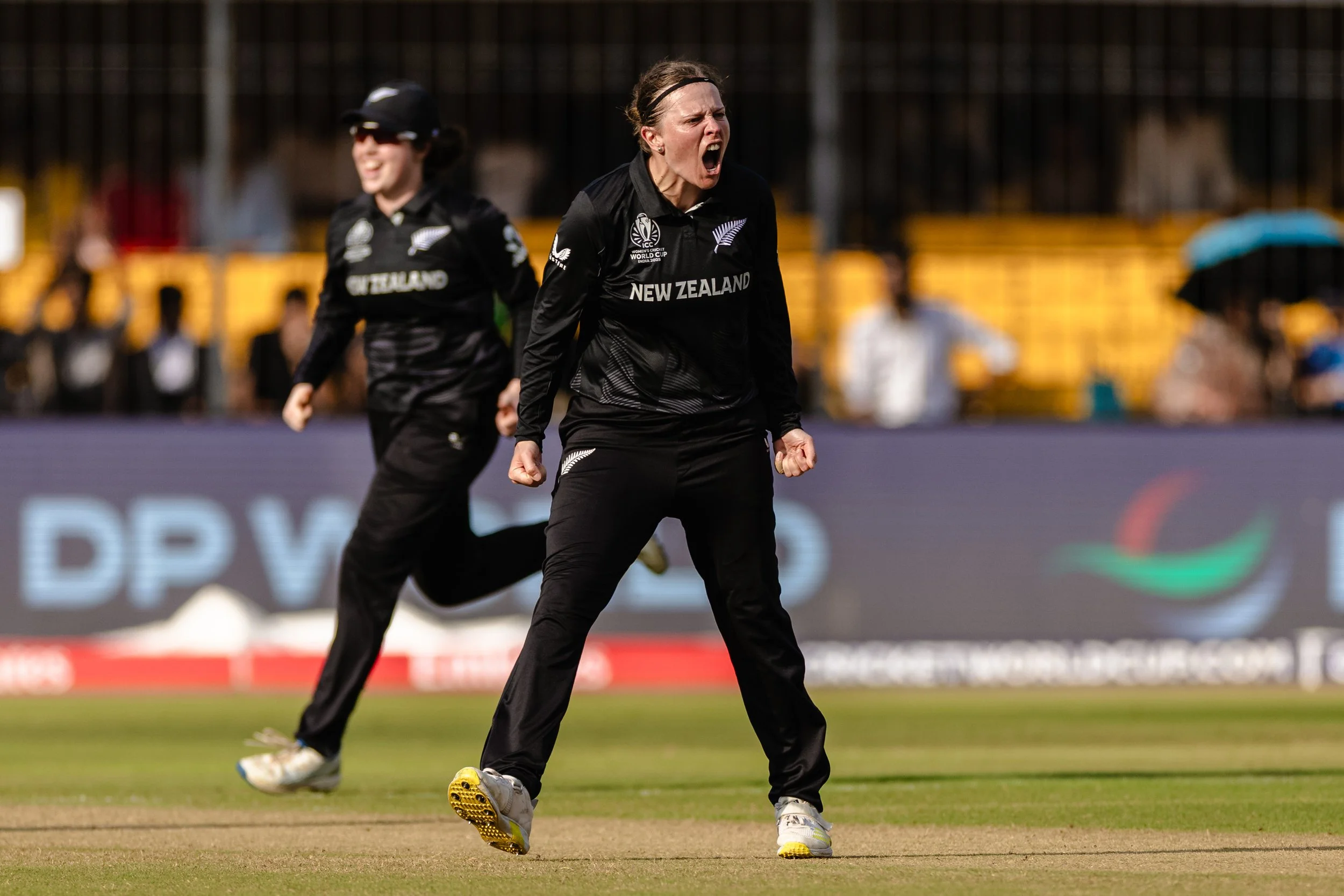 A woman from New Zealand cricket team celebrating during a match, with a woman in the background also in team uniform, on the cricket field.