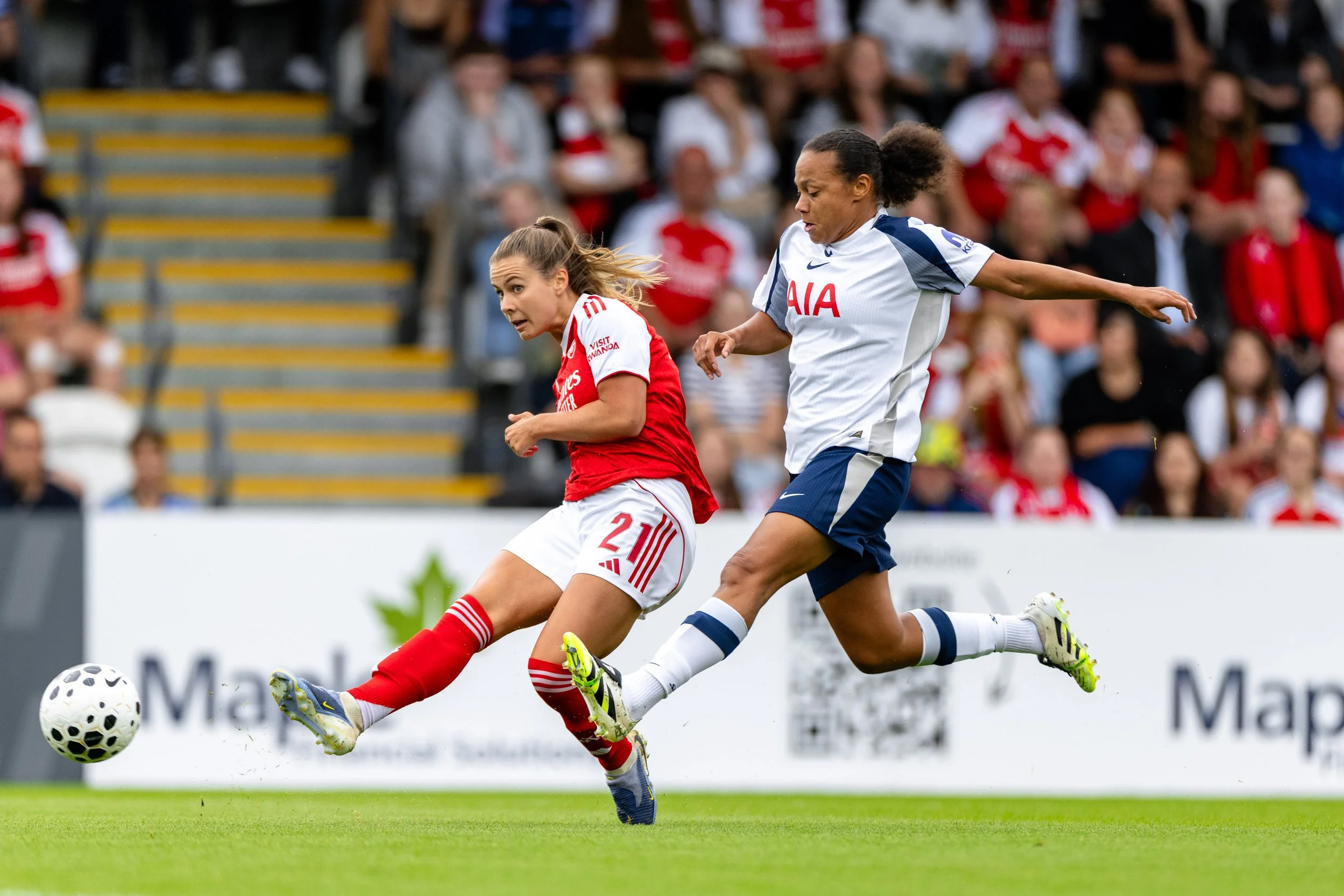 Two female soccer players competing for the ball during a match, with one in a red and white uniform and the other in a white and navy uniform, on a green field with spectators in the background.