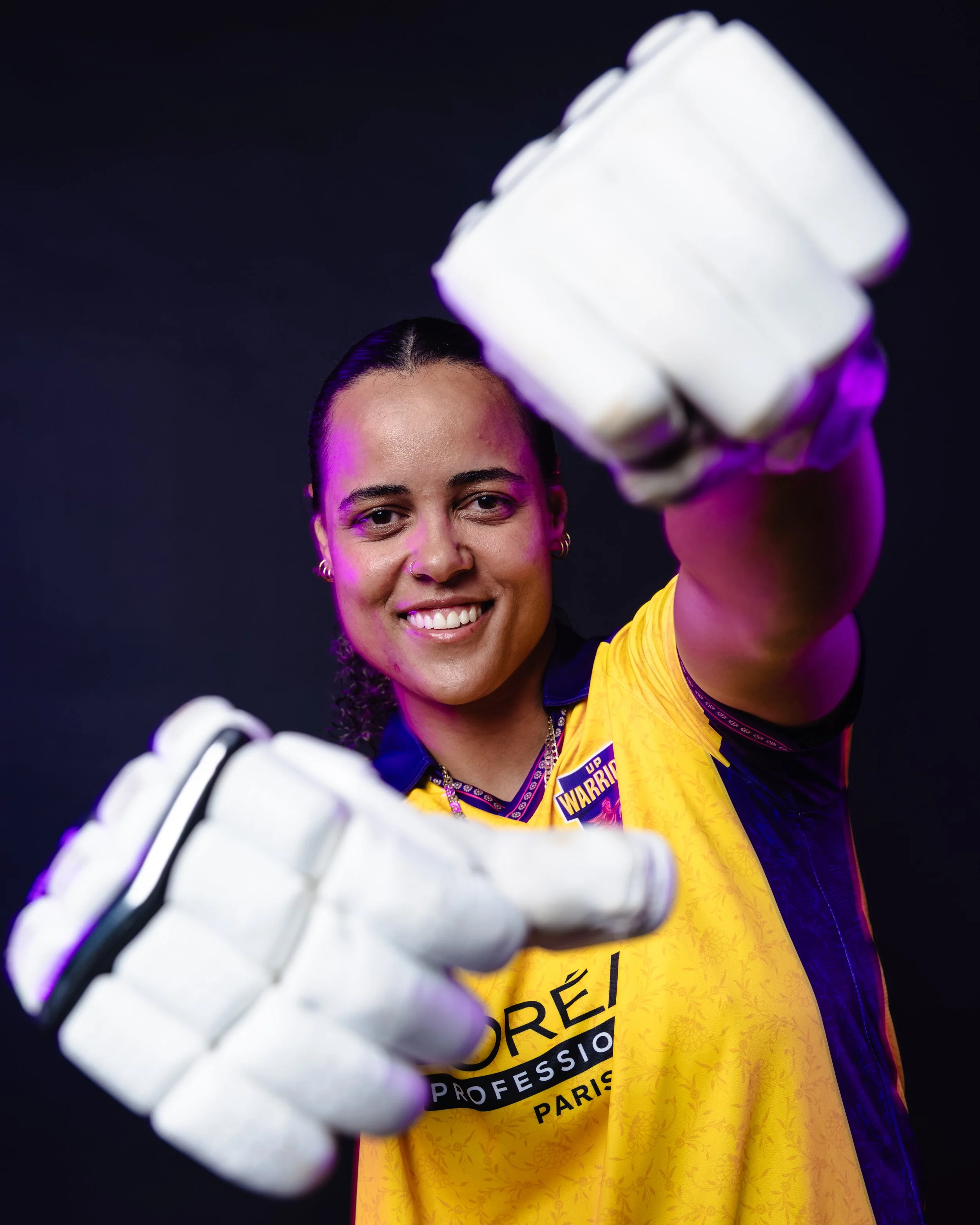 Close-up of a young woman with dark hair, smiling, wearing a yellow sports jersey with black lettering, and a purple and black jacket, holding a cricket helmet towards the camera with a black background.