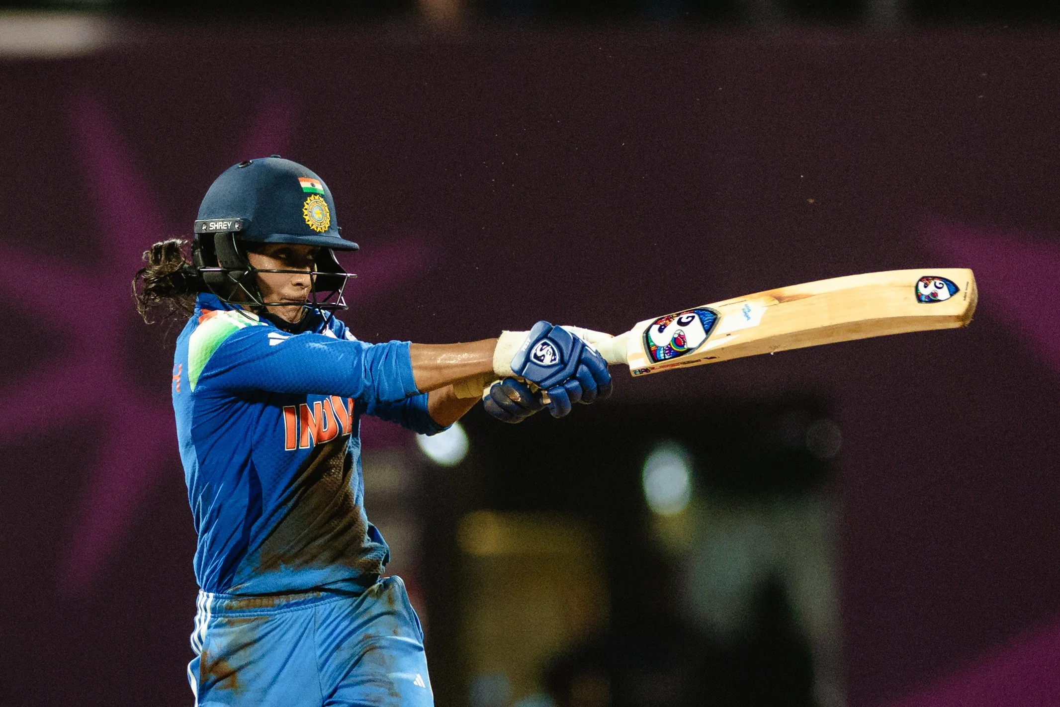 A female cricket player wearing a blue jersey, helmet, and gloves swinging a wooden cricket bat during a match.