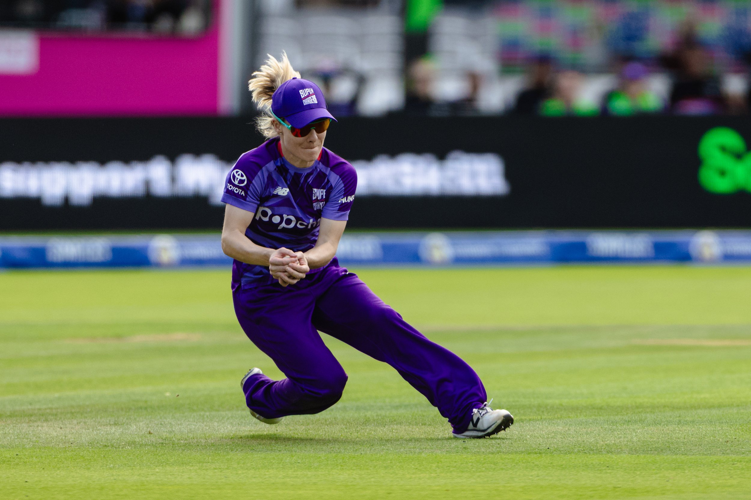 A female cricket player in purple uniform with a purple cap and sunglasses on the field during a match.
