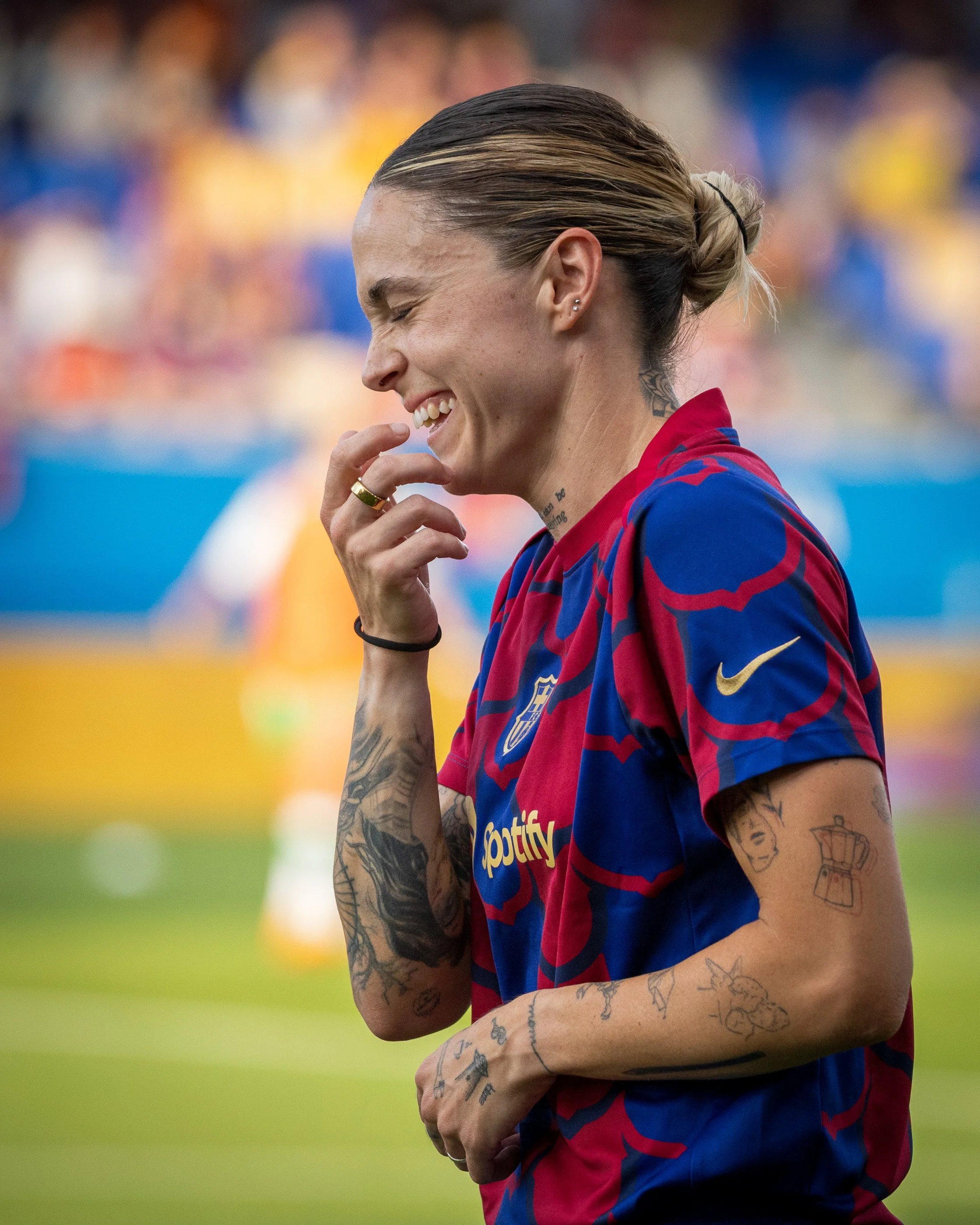 A female soccer player wearing a red and blue jersey with tattoos on her arms and neck, smiling with eyes closed and hand near her face, on a soccer field.