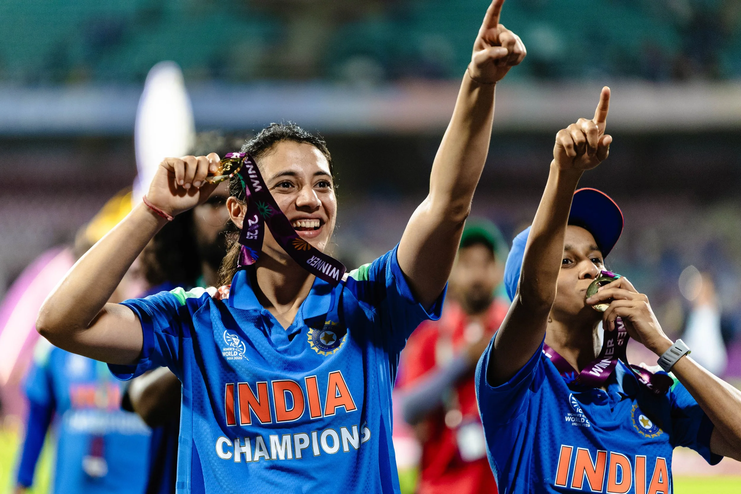 A group of women wearing blue cricket jerseys, smiling, and celebrating after winning a match. One woman is holding a medal and the team jerseys display 'India' and 'Champions'.