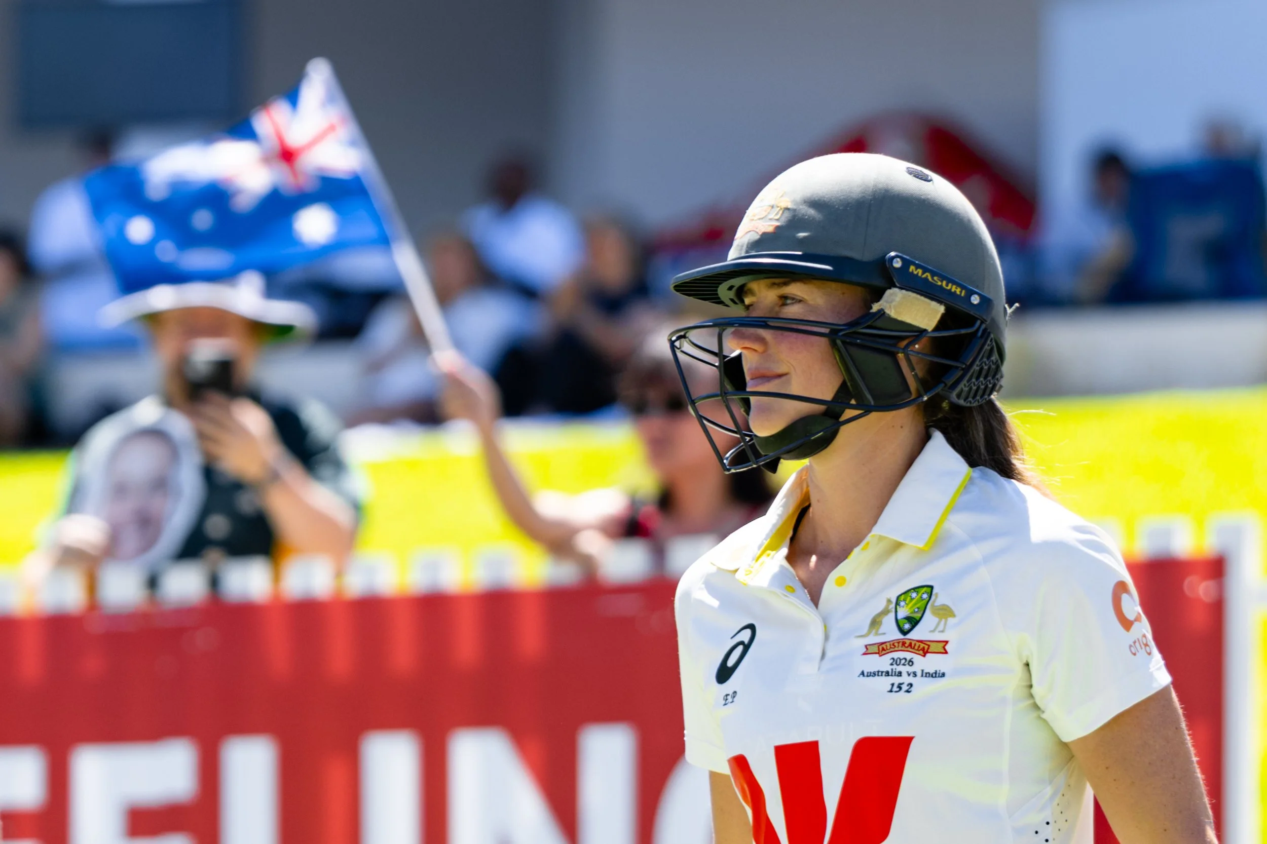 A female cricket player wearing a helmet and white jersey during a match, with Australian colors and patches, with spectators and a flag in the background.