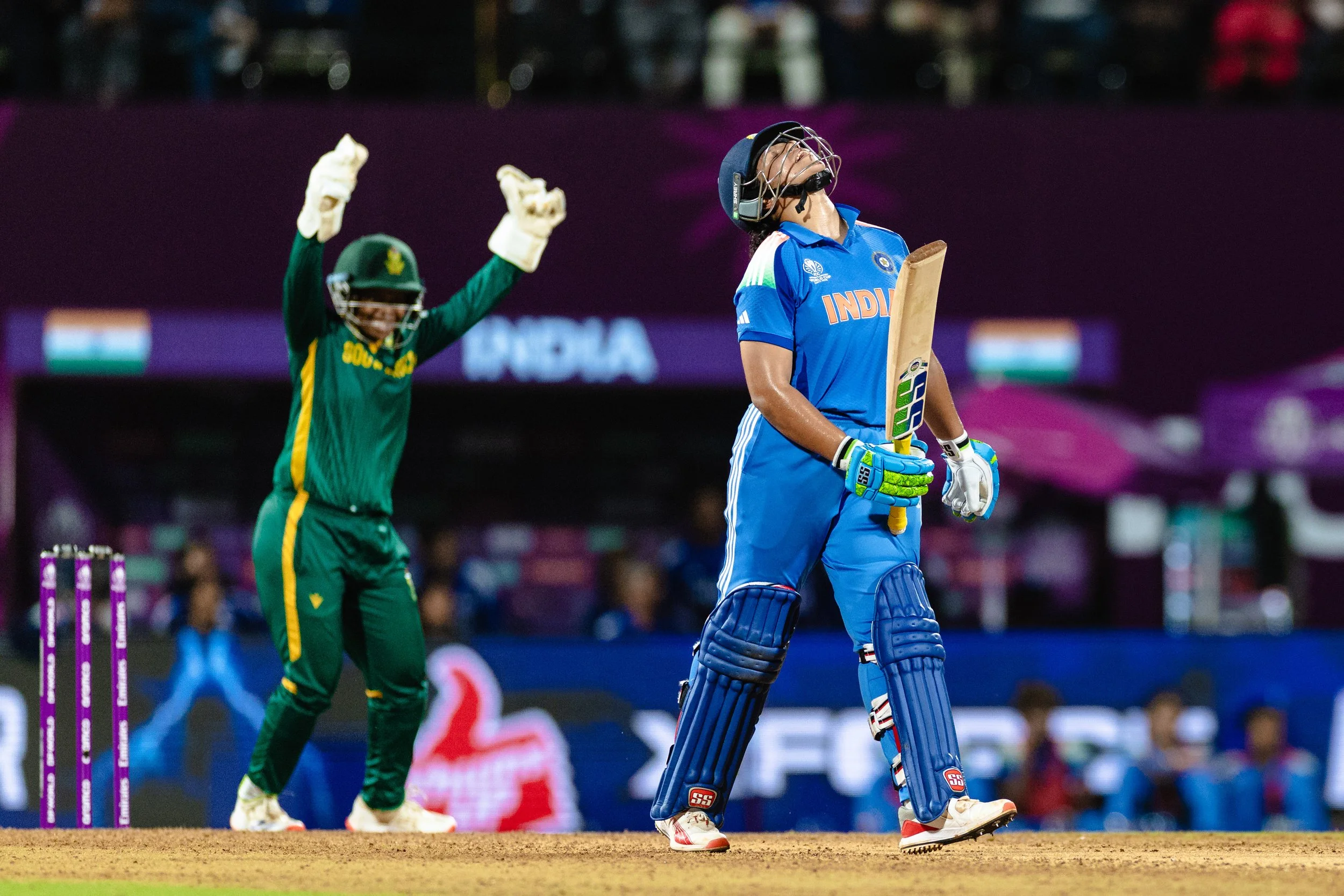 An Indian female cricket player in blue protective gear and gloves, holding a cricket bat, looks up with a relieved expression. An South African male wicketkeeper in green and yellow gear celebrates with raised arms in the background, during a cricke