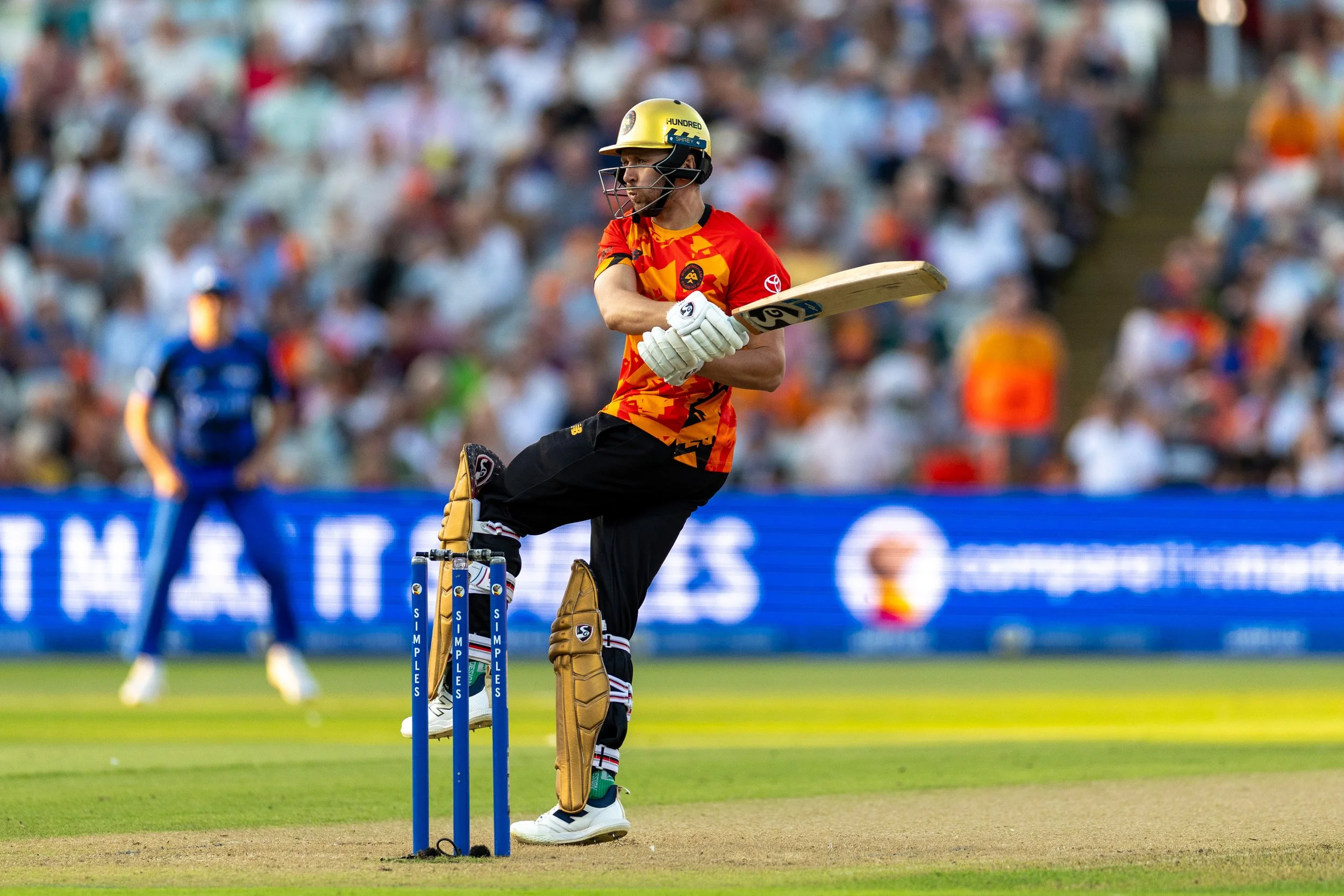 A cricket player wearing protective gear and a colorful red and yellow jersey is taking a stance on the field during a match, with a crowd of spectators blurred in the background.