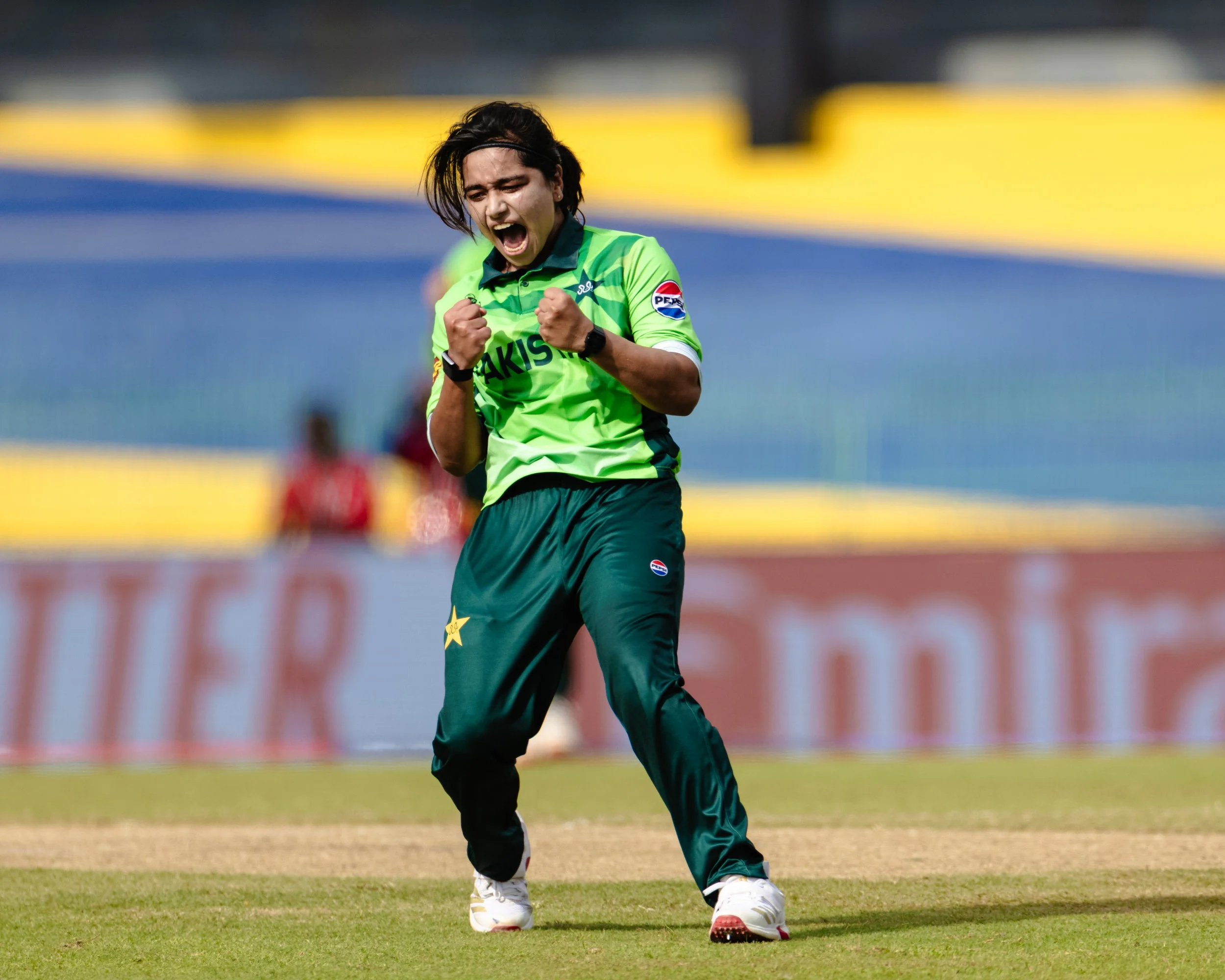 A woman wearing a bright green sports uniform celebrating with fists clenched in front of a sports field.