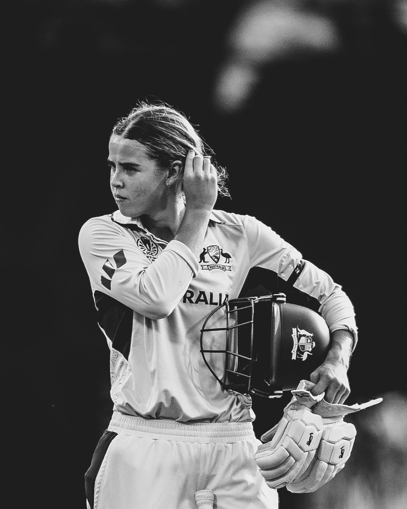 A female cricket player in a uniform holding a helmet and a piece of paper, adjusting her hair with one hand.