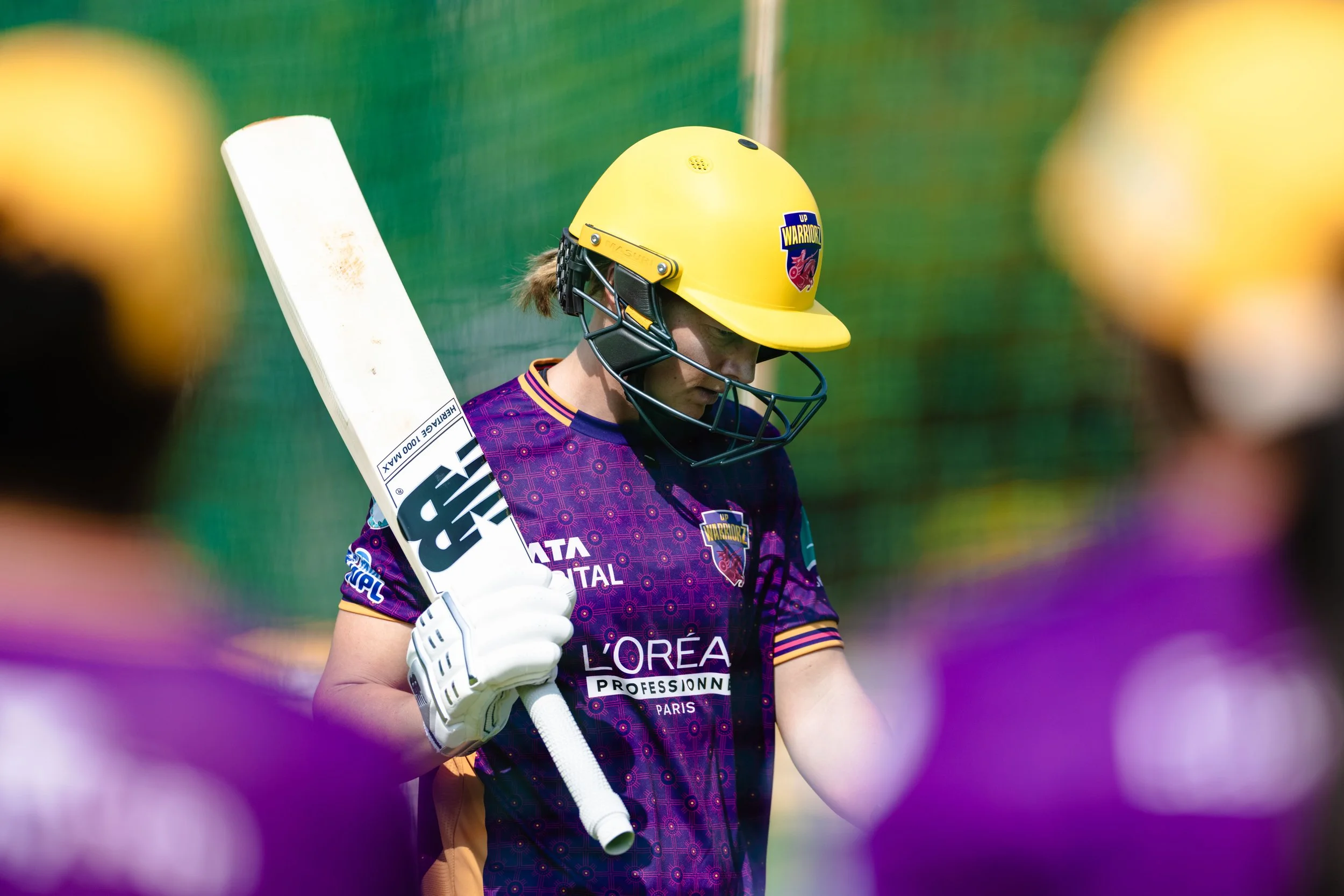 A female cricket player wearing a yellow helmet with a logo, purple jersey, and white gloves, holding a cricket bat during a game.