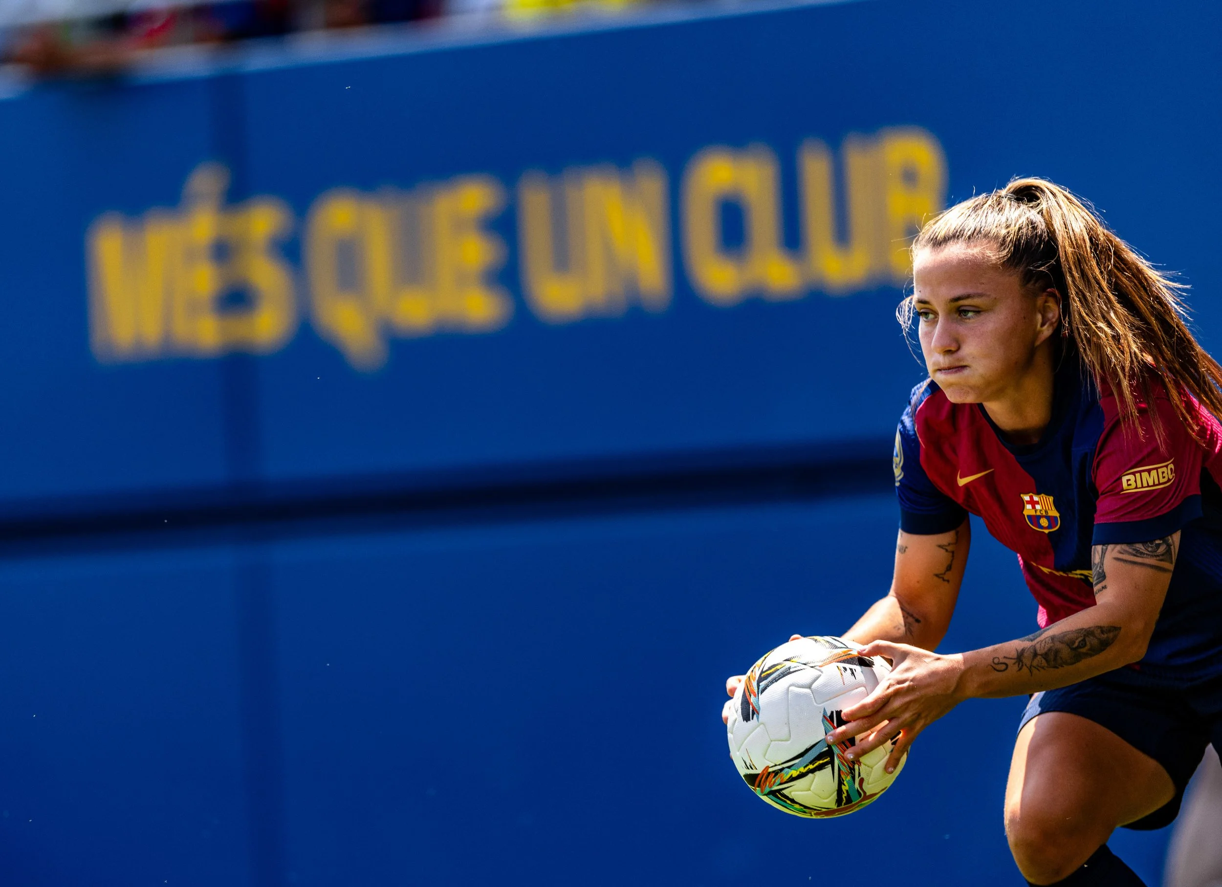 A female soccer player with tattoos on her arm, wearing a red and blue jersey, is holding a white soccer ball with colorful designs on it on a blue field.