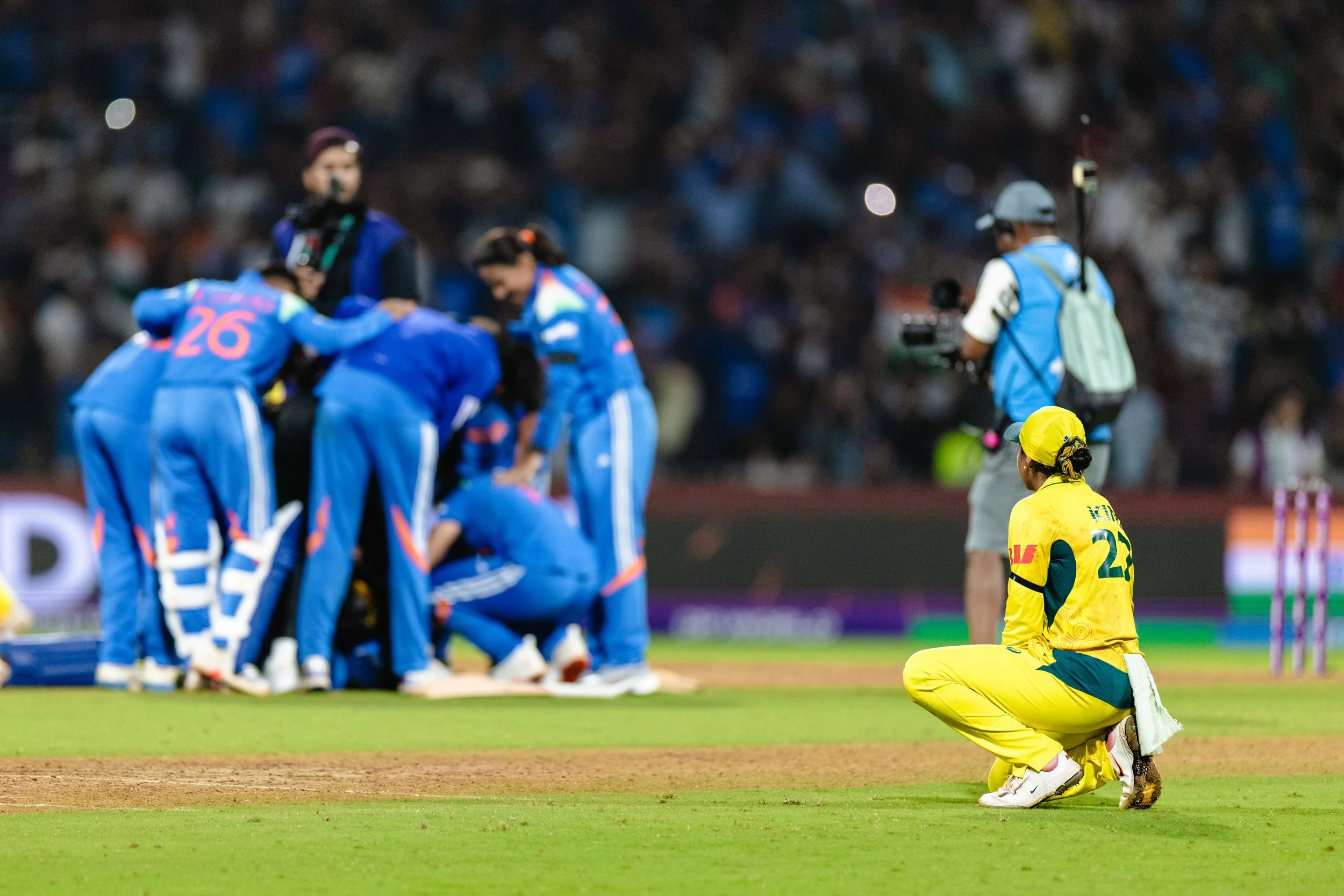 Kneeling cricket player in yellow uniform on the pitch during a match, with a cluster of players in blue uniforms celebrating in the background in a stadium setting.