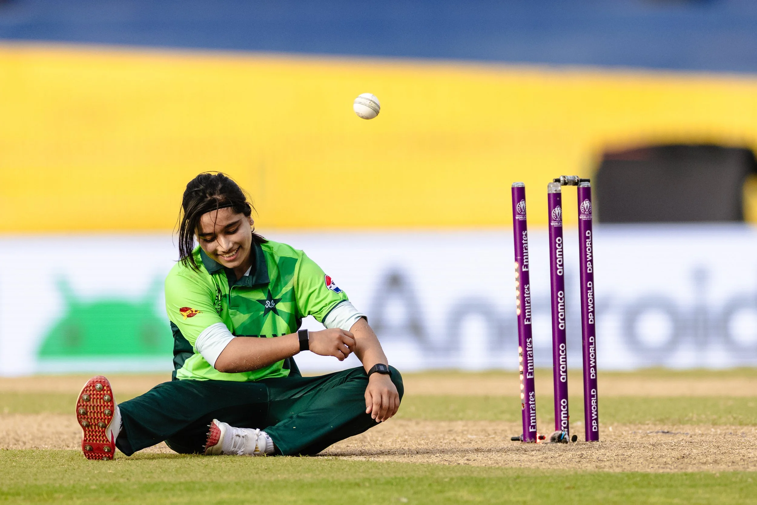 A female cricket player sitting on the ground after an attempt at a shot, with wickets in the background during a match.