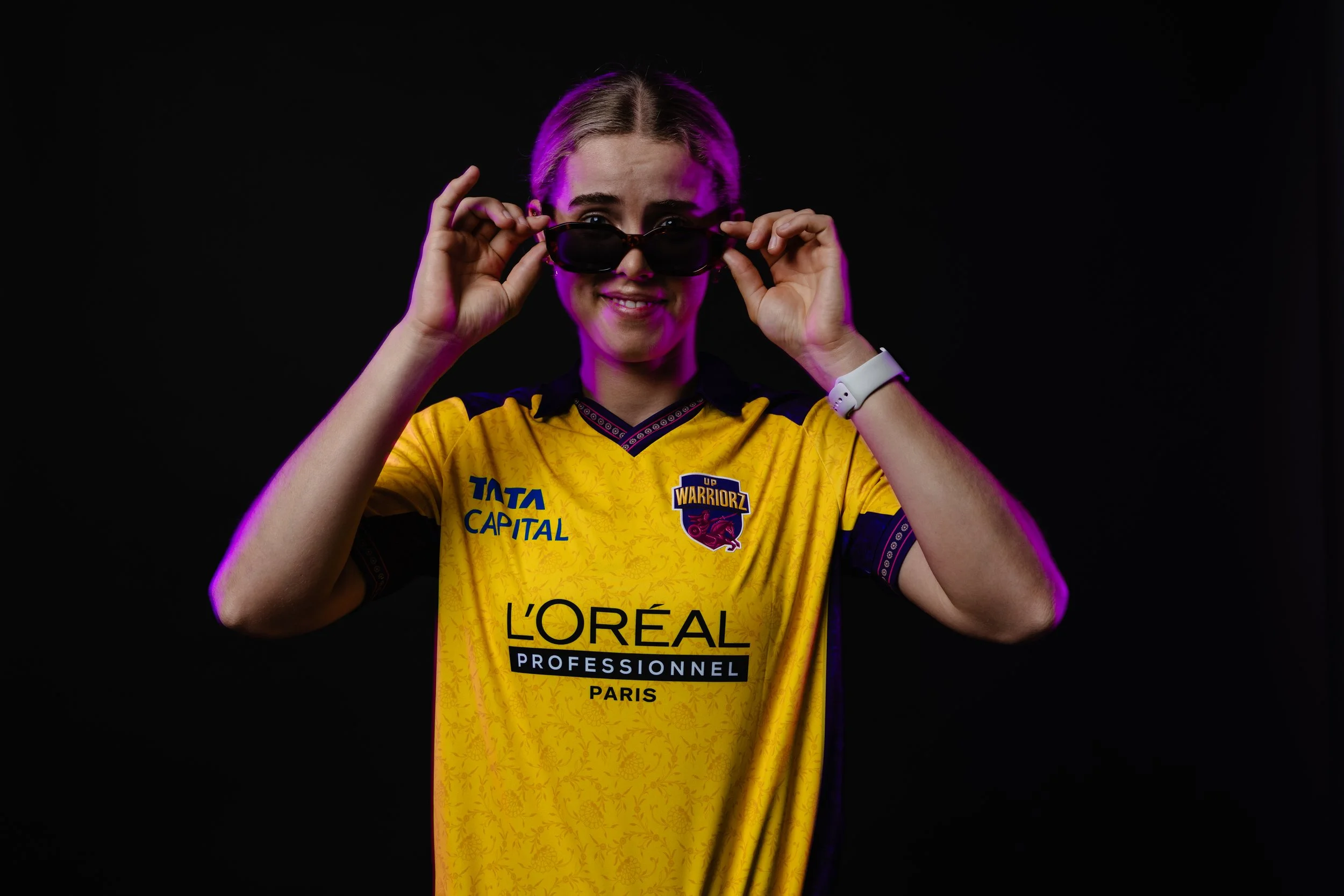 A young woman with blonde hair wearing a yellow sports jersey and sunglasses, smiling while looking at the camera against a black background.