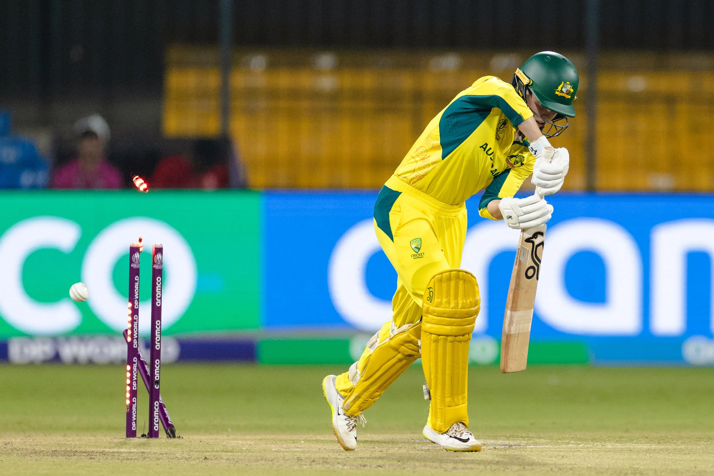 Cricket player in yellow and green uniform with protective gear, bending down to play a shot during a match with stumps and floodlights in the background.