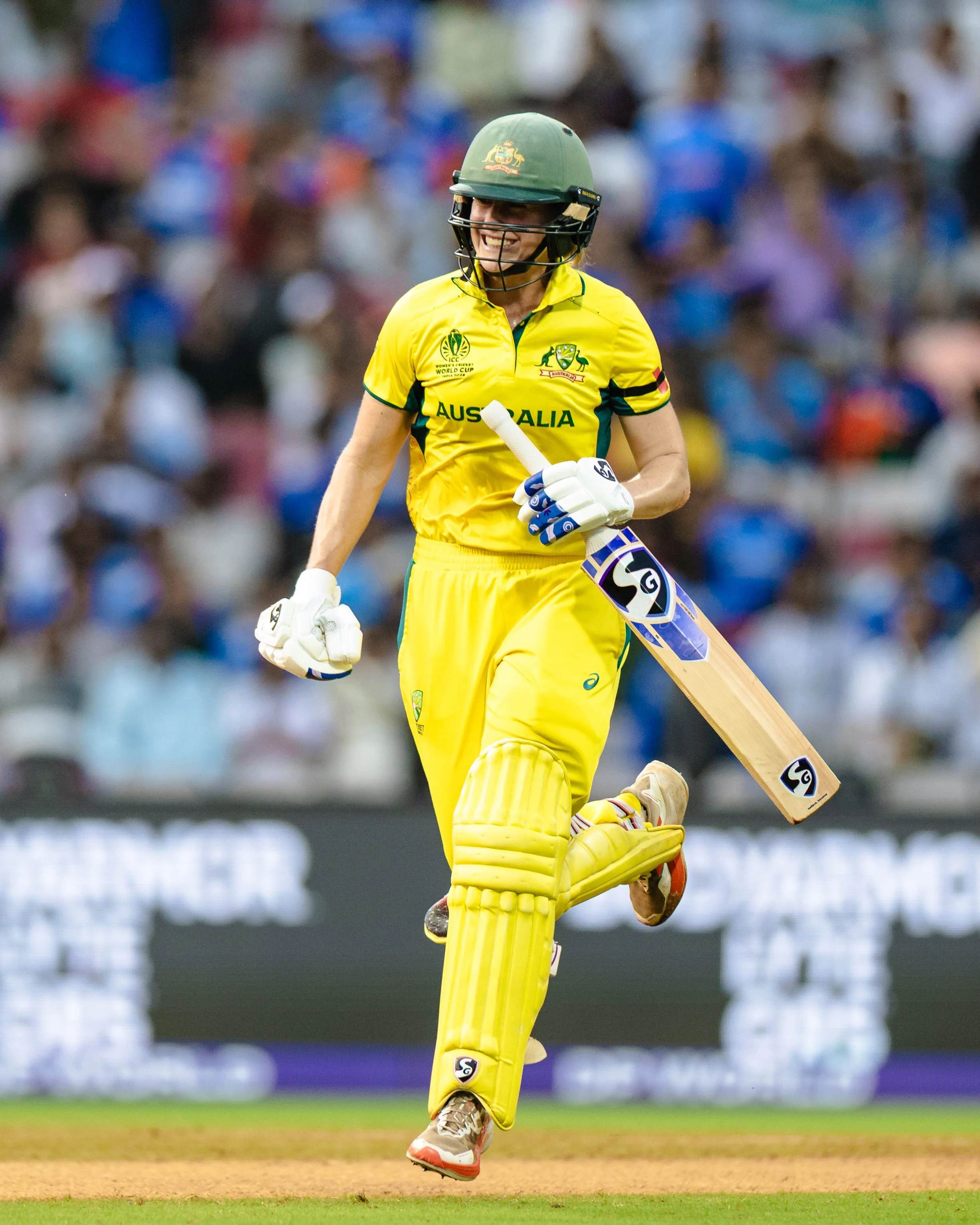 Cricket player in yellow uniform celebrating on the field with a smile, holding a cricket bat, with a blurred crowd in the background.