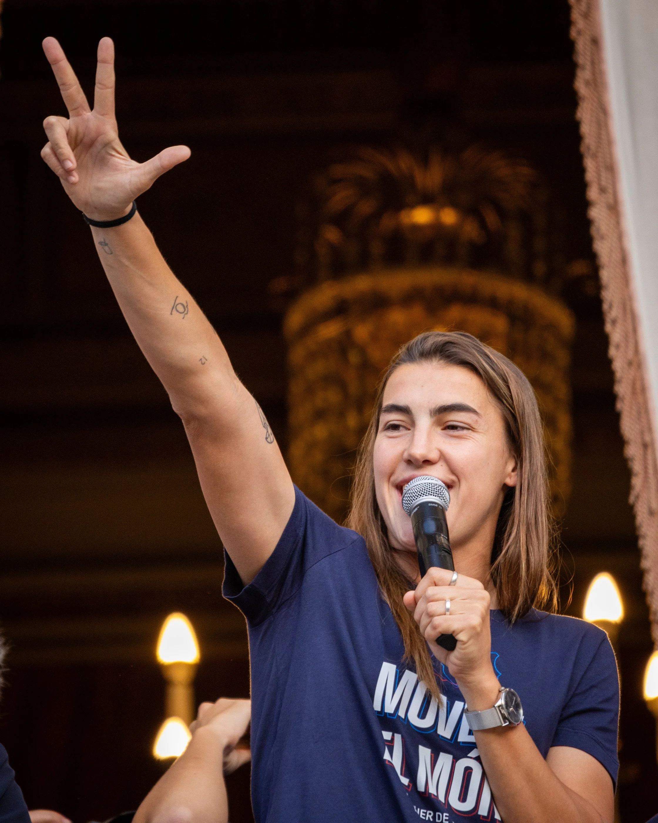 A woman with light brown hair, smiling and speaking into a microphone, raising her right hand in a sign of victory or peace, with a dark background and warm lighting.