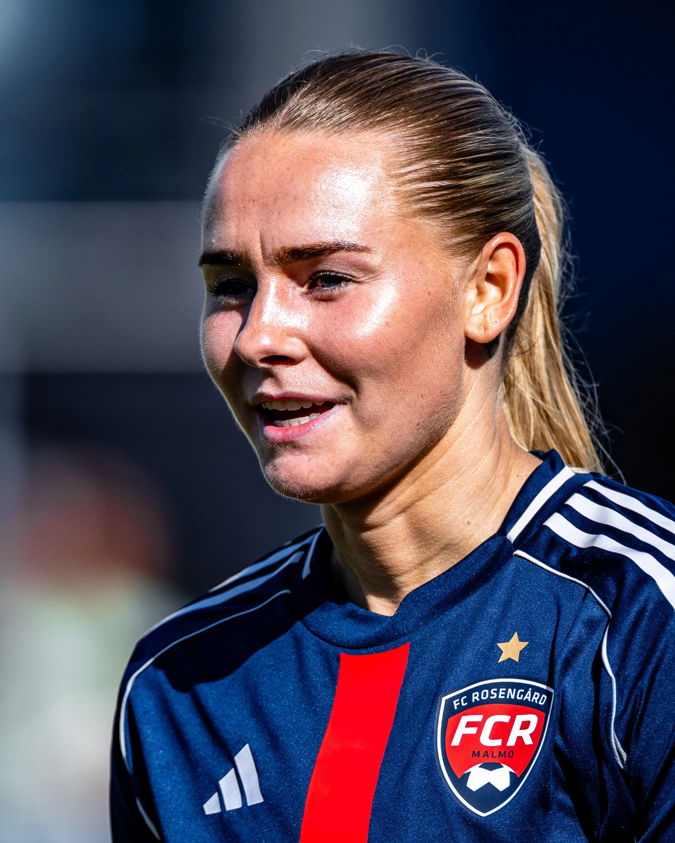 Close-up of a woman with blonde hair tied back, wearing a blue sports jersey with stripes and a crest that reads 'FC Rosengård Malmö', on a soccer field.
