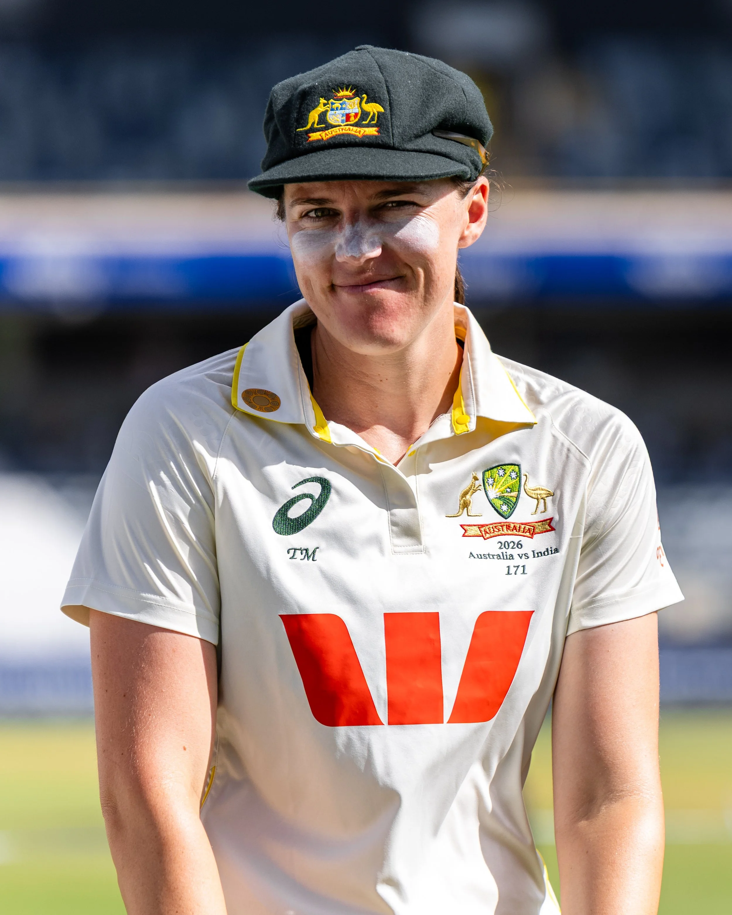 Close-up of a person's upper body in cricket attire, wearing a white shirt with yellow accents and a crest that indicates Australia. The person has a black hat with an Australian emblem and face paint on their nose. The background is blurred with blu