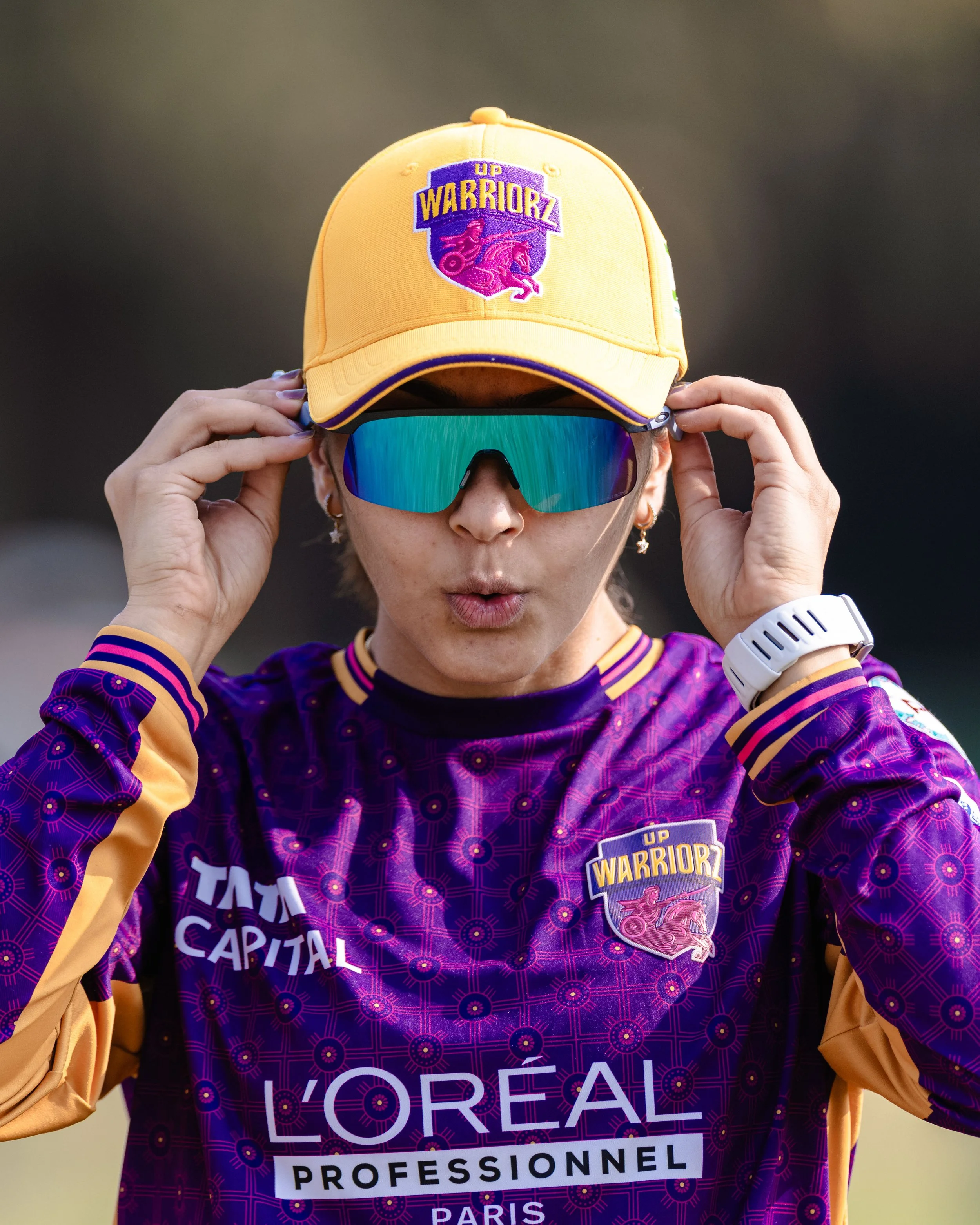 A woman wearing sunglasses, a purple sports jersey with gold and purple accents, a yellow cap with an embroidered logo that reads 'UP WARRIORZ,' and a white wristwatch. The jersey displays several sponsor logos including L'Oréal Professionnel and oth