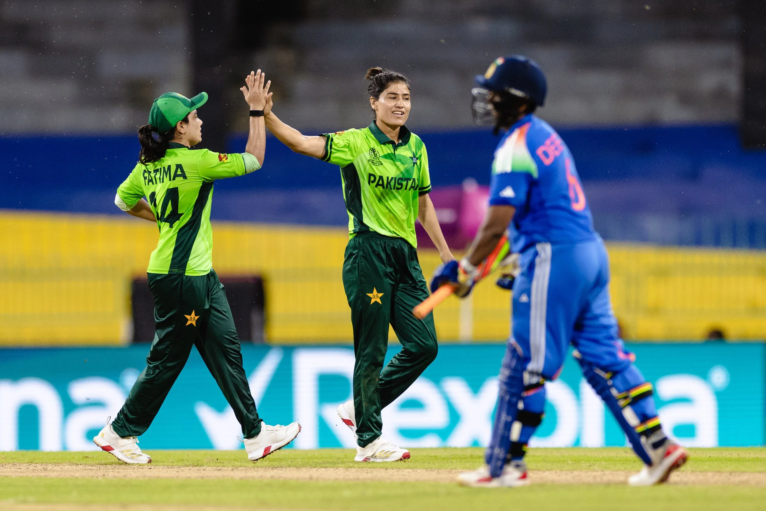 Two female cricket players with green uniforms from Pakistan high-five, while an opposing female player in a blue uniform watches, on the field during a match.